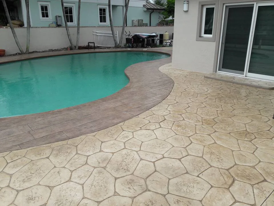 Backyard pool deck area with stone textured decorative stamped concrete patio and decorative concrete path along the pool with wooden stamped concrete detail and colors adjacent to a house with sliding glass doors.
