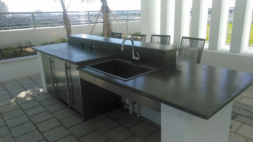 Outdoor kitchen with gray decorative concrete countertops, stainless steel sink, faucet, and cabinets on a tiled patio.