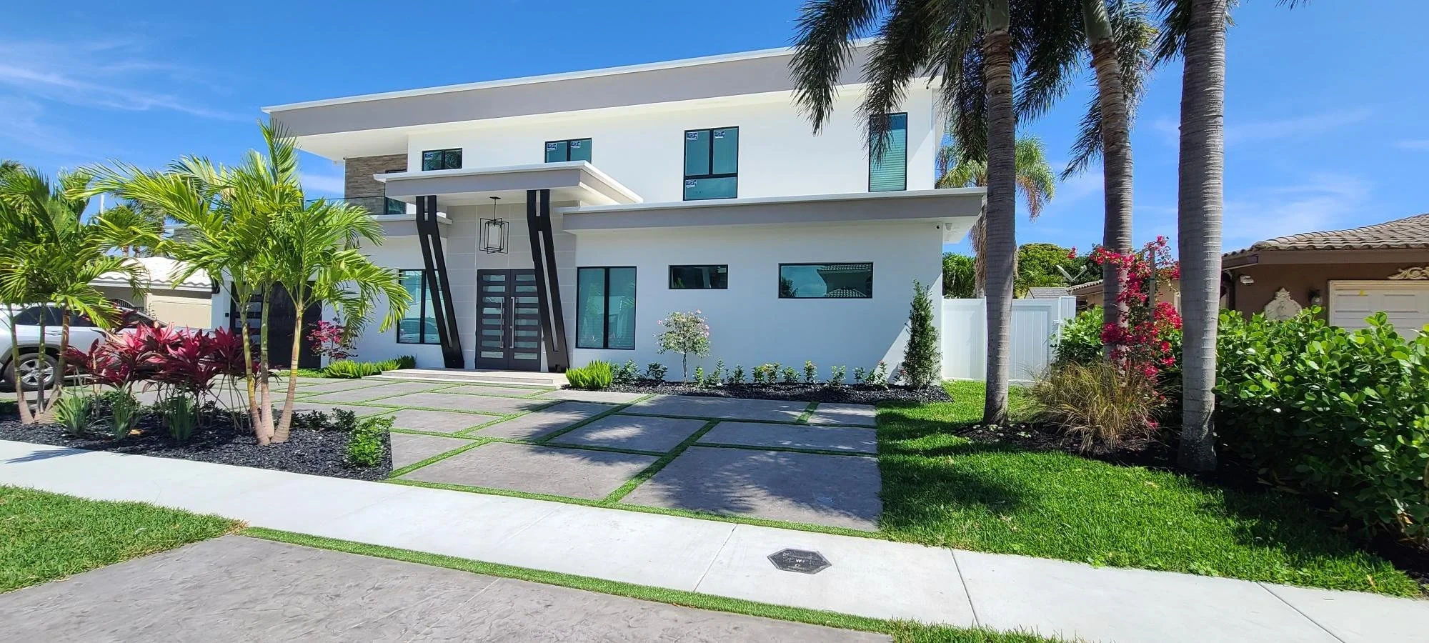 Modern white two-story house with large windows, surrounded by palm trees and landscaped garden.