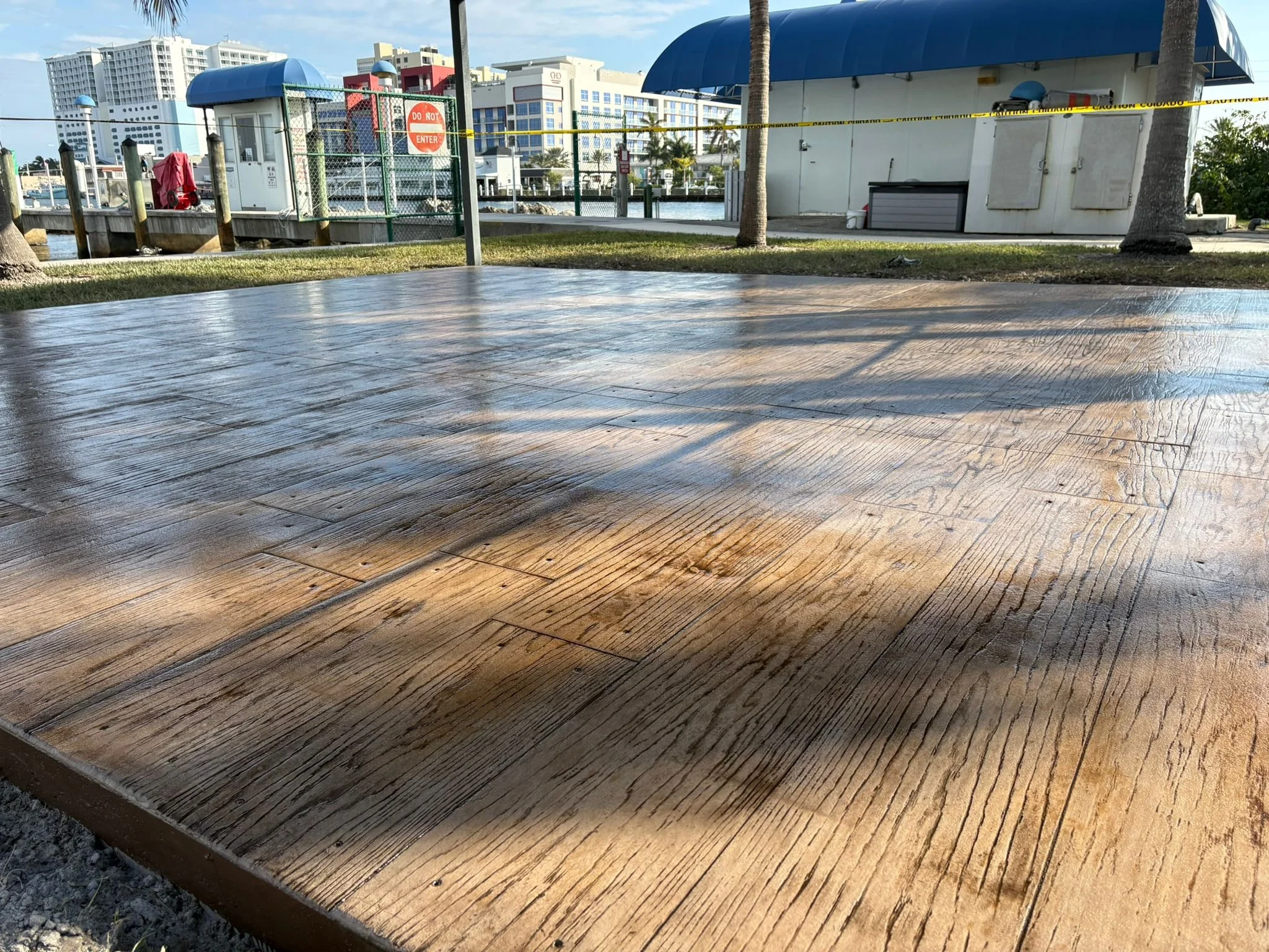 Wet concrete slab with wood texture pattern near waterfront, surrounded by buildings and palm trees.