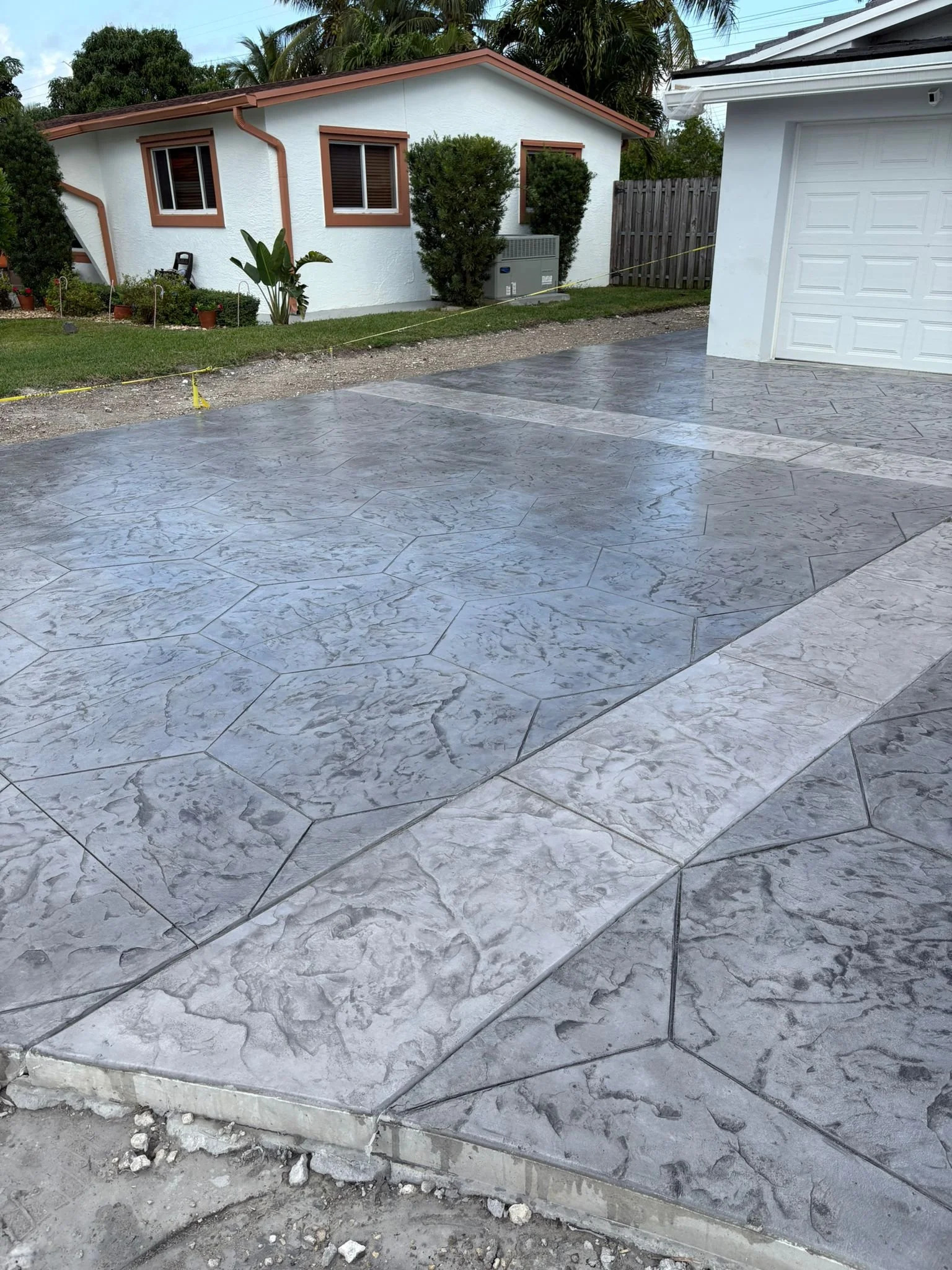 Textured stamped concrete driveway in front of a white house with brown trim, surrounded by greenery.