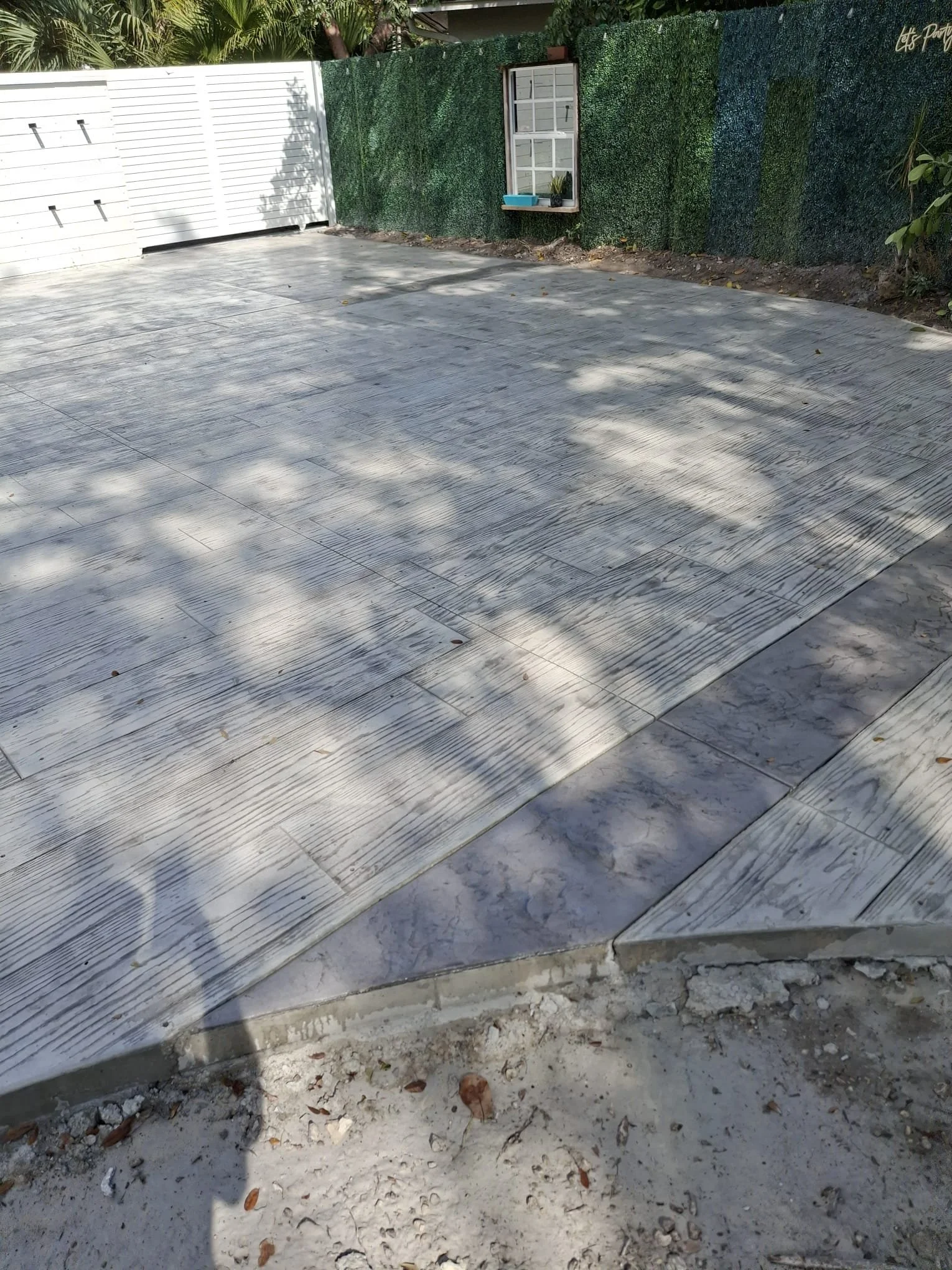Concrete patio with textured surface, bordered by artificial green hedge and a small window with a planter.