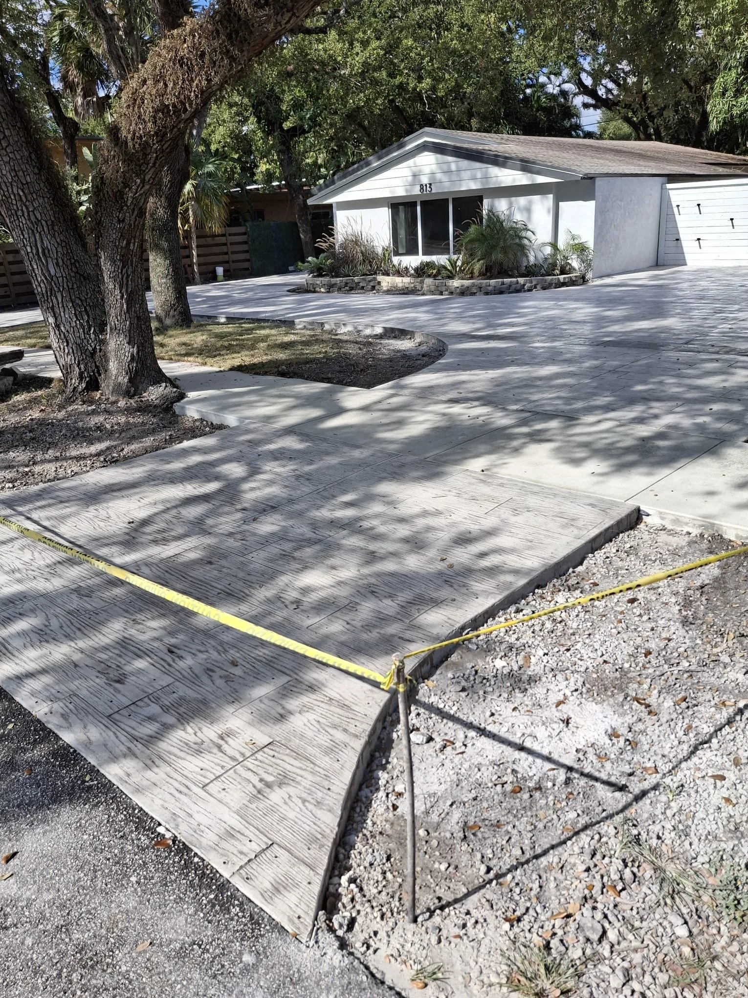 Residential driveway under construction with new concrete, surrounded by yellow caution tape, leading to a single-story white house with a garage, trees, and landscaping in the front yard.