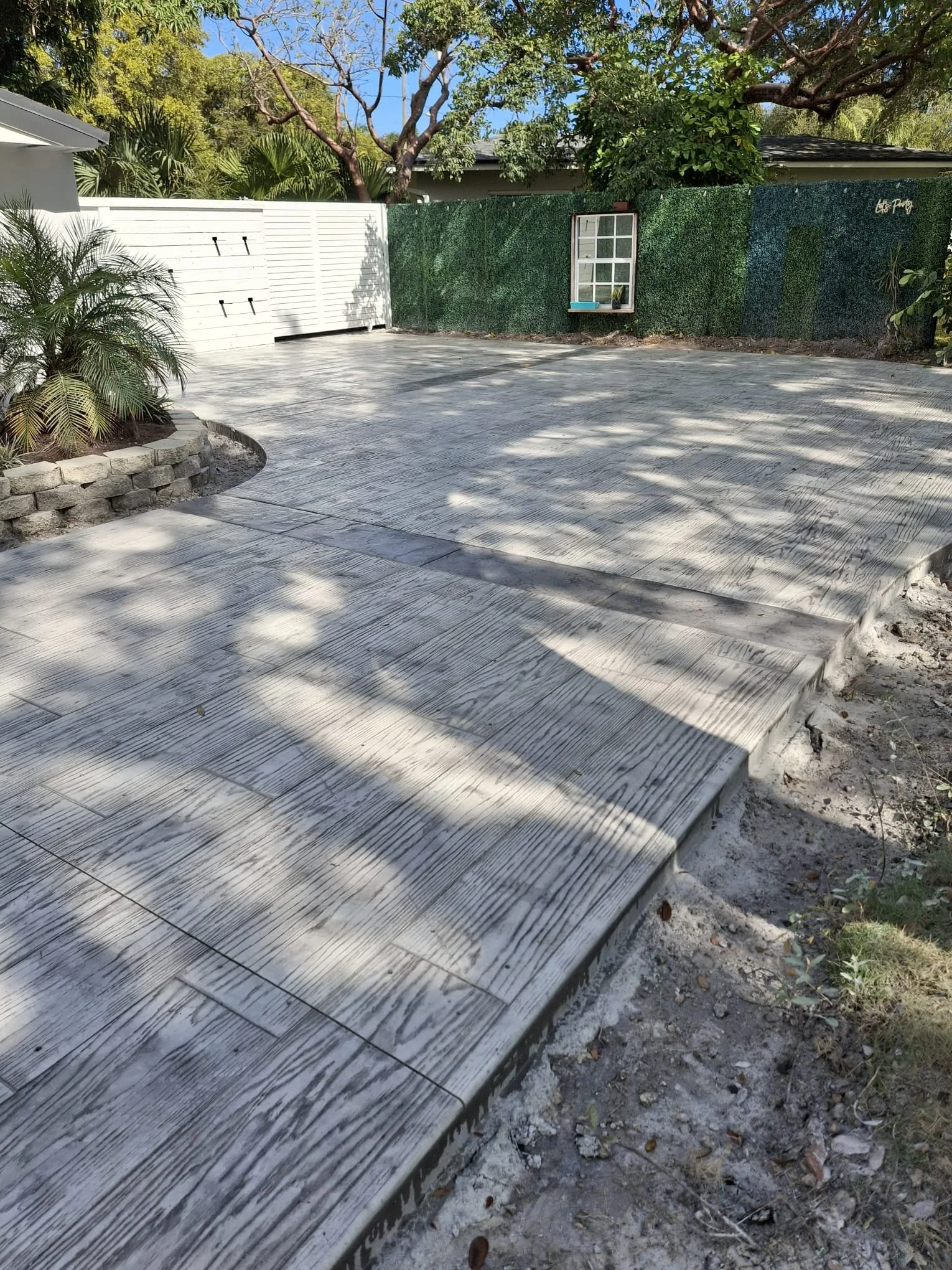 A backyard with a concrete patio displaying a wood-like texture, surrounded by a white fence, green foliage, and trees.