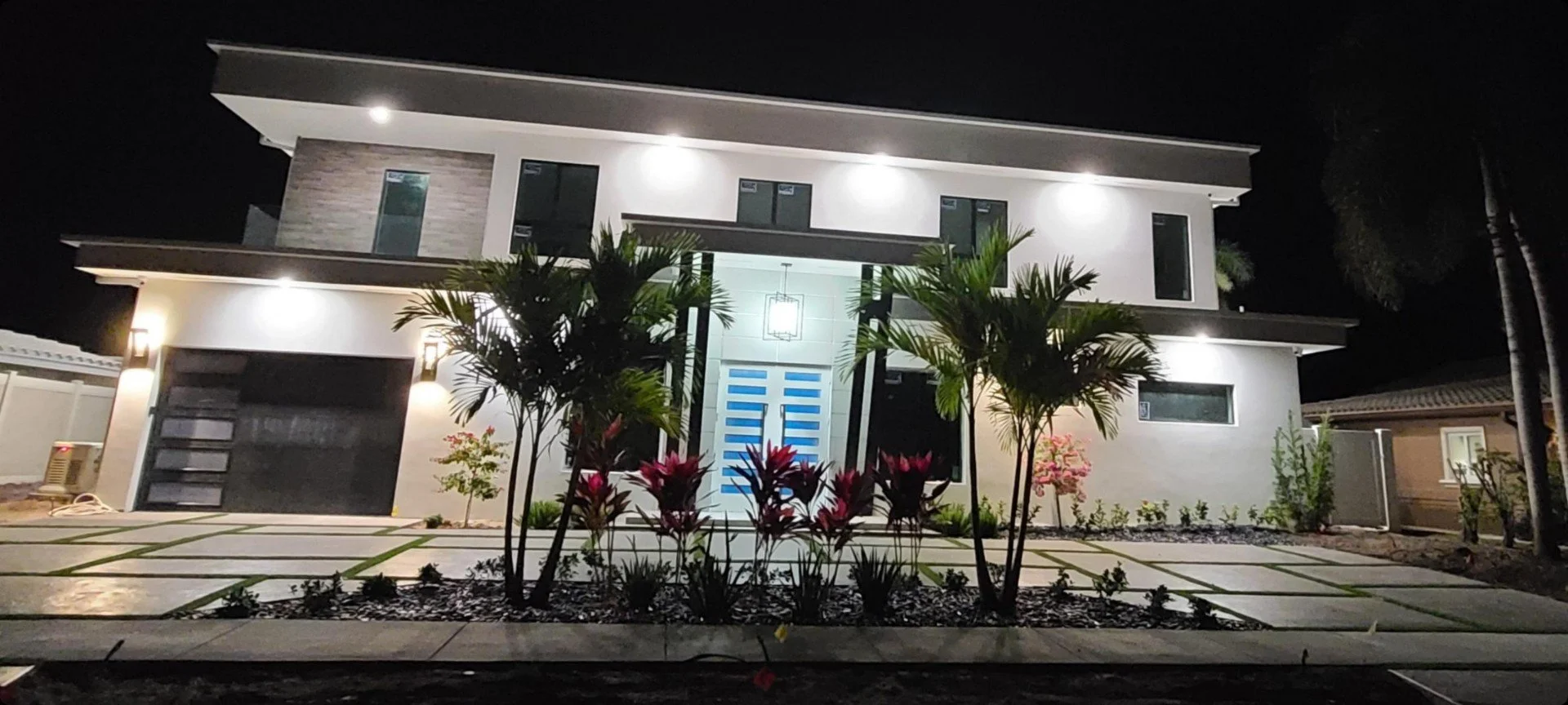 Designed Stamped Concrete drive way on a modern two-story house with large glass windows, illuminated at night, surrounded by palm trees and landscaped front yard.