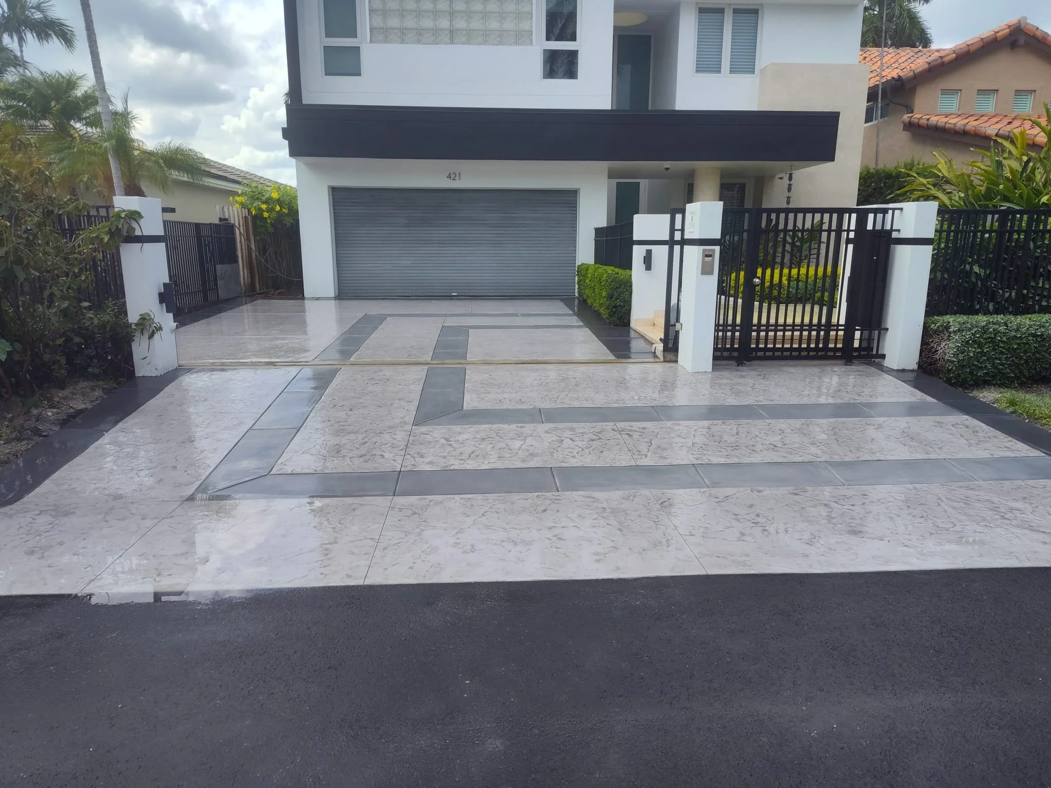 Modern two-story house with a patterned driveway, gray garage door, and surrounding plants and trees.
