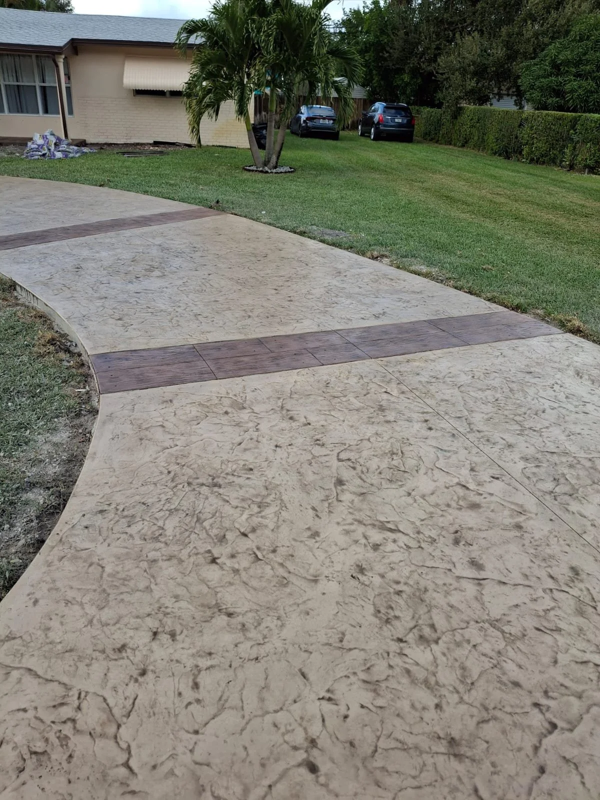 Residential driveway with stamped concrete texture, bordered by green grass and a palm tree, leading to a beige house with awnings and parked cars.