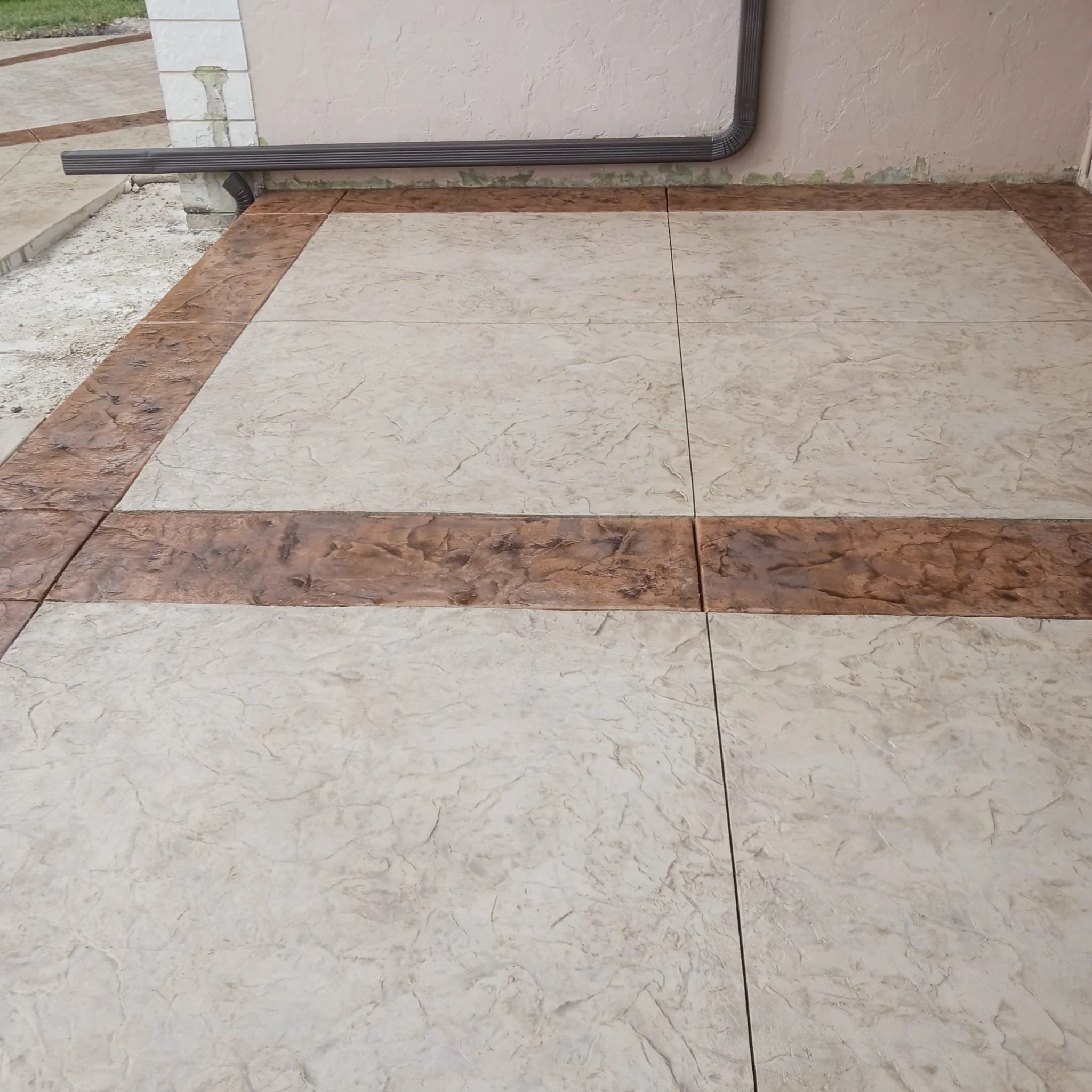 Textured concrete patio with light tan tiles bordered by darker brown tiles.
