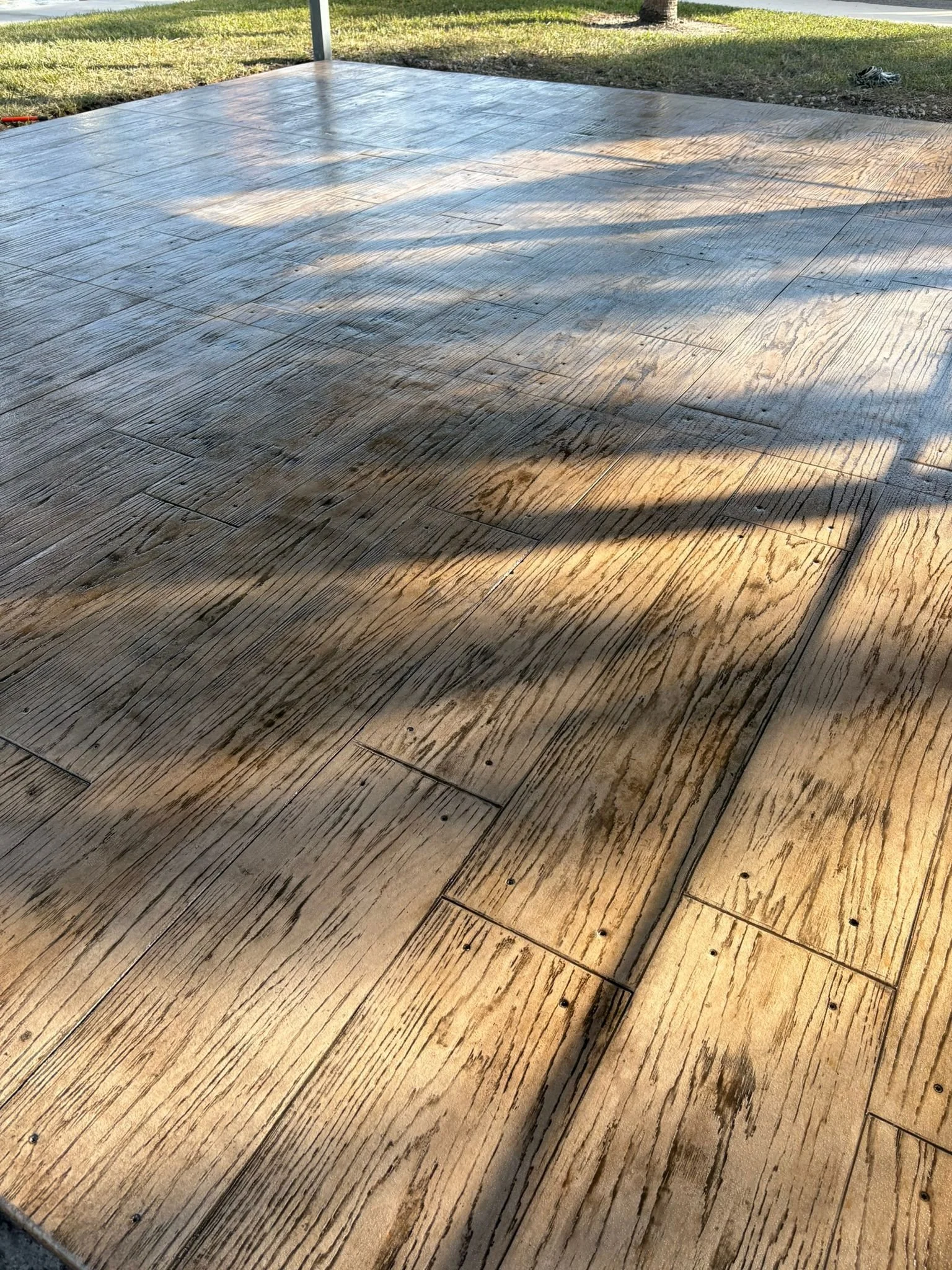 Concrete patio with wood-like stamped texture, showing sunlit patterns, surrounded by grass.