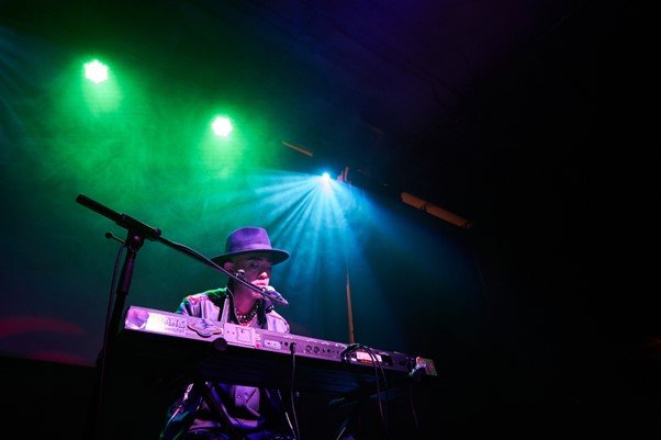 The Mollusc Dimension is an East Asian artist on stage playing the keyboard with a mic stand in front of him and dramatic green and blue haze lighting streaming down from above.