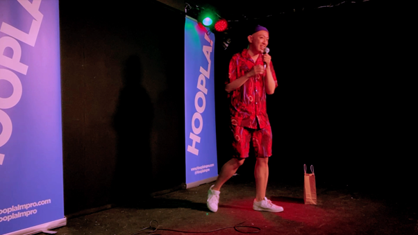The Mollusc Dimension, an East Asian transqueerian, is on stage holding a mic and performing stand-up. He looks happy and bouncy and wears a flamboyant red floral shorts set and white trainers.