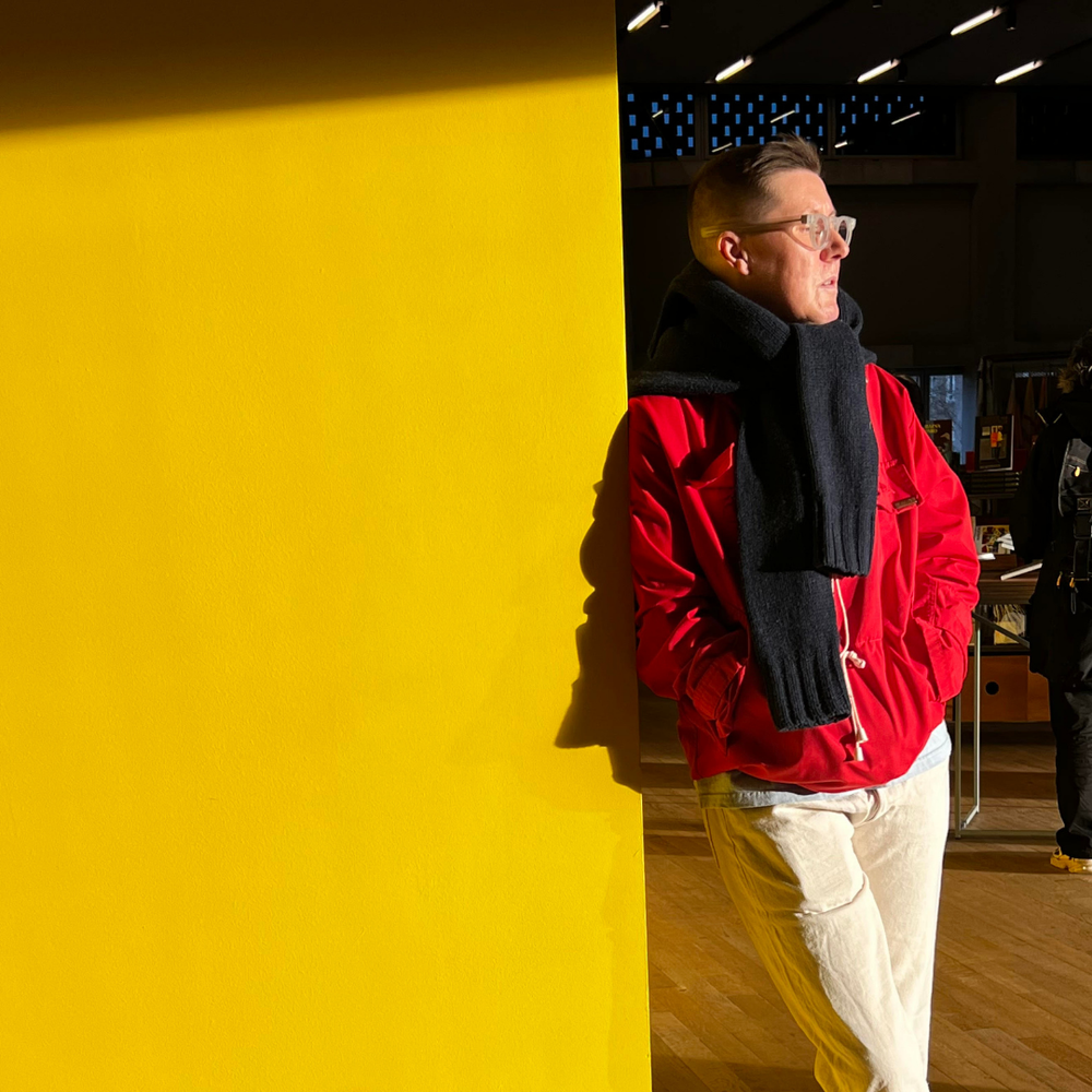 Tink, a white masculine person, leaning against a bright yellow wall
