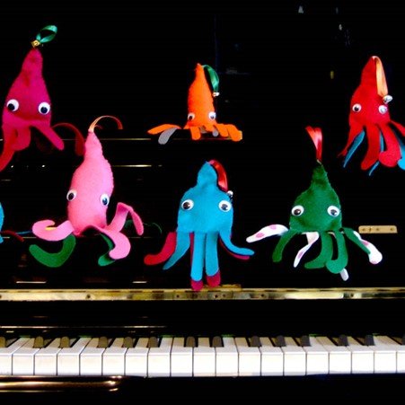 6 felt squids in bright colours sit in an adorable yet declamatory formation on the lid and music stand of a black upright piano above the keyboard.  The squids each have a ribbon coming from their mantle and googly eyes. Some have bells attached.