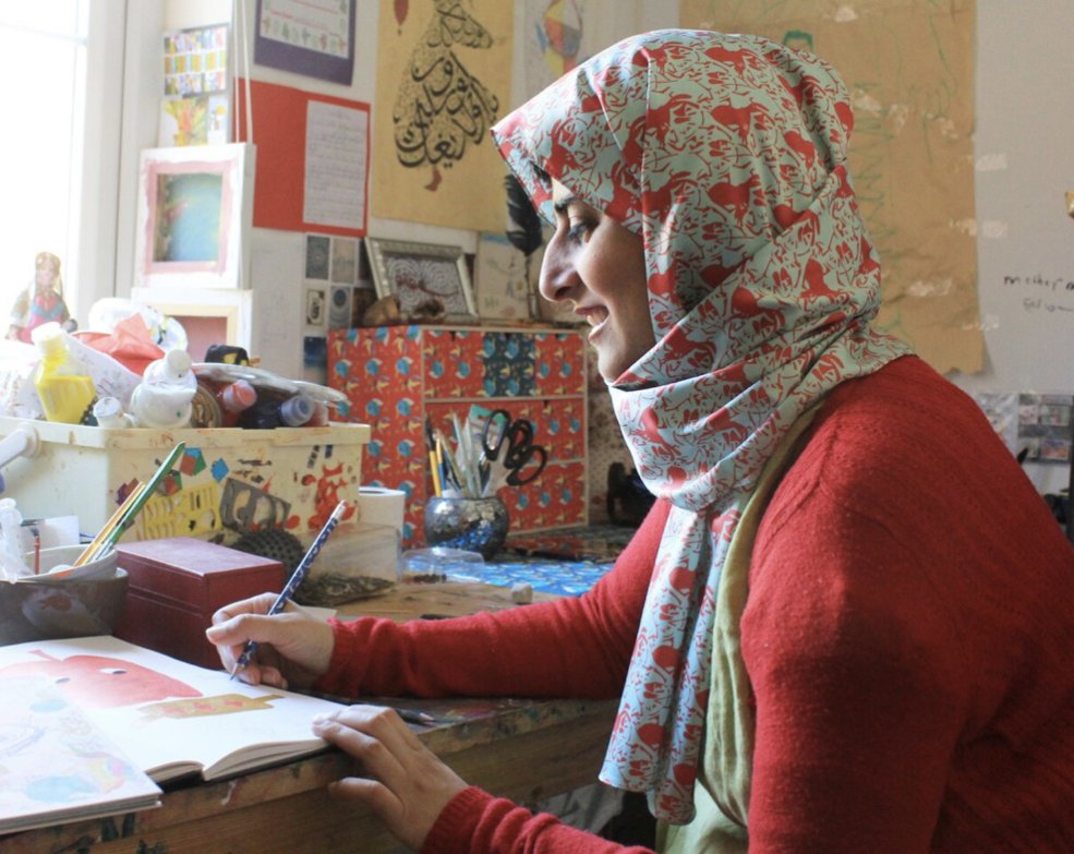 A woman wearing a patterned headscarf and red sweater sitting at a cluttered desk, drawing in a sketchbook with art supplies around her, in an art studio or classroom.