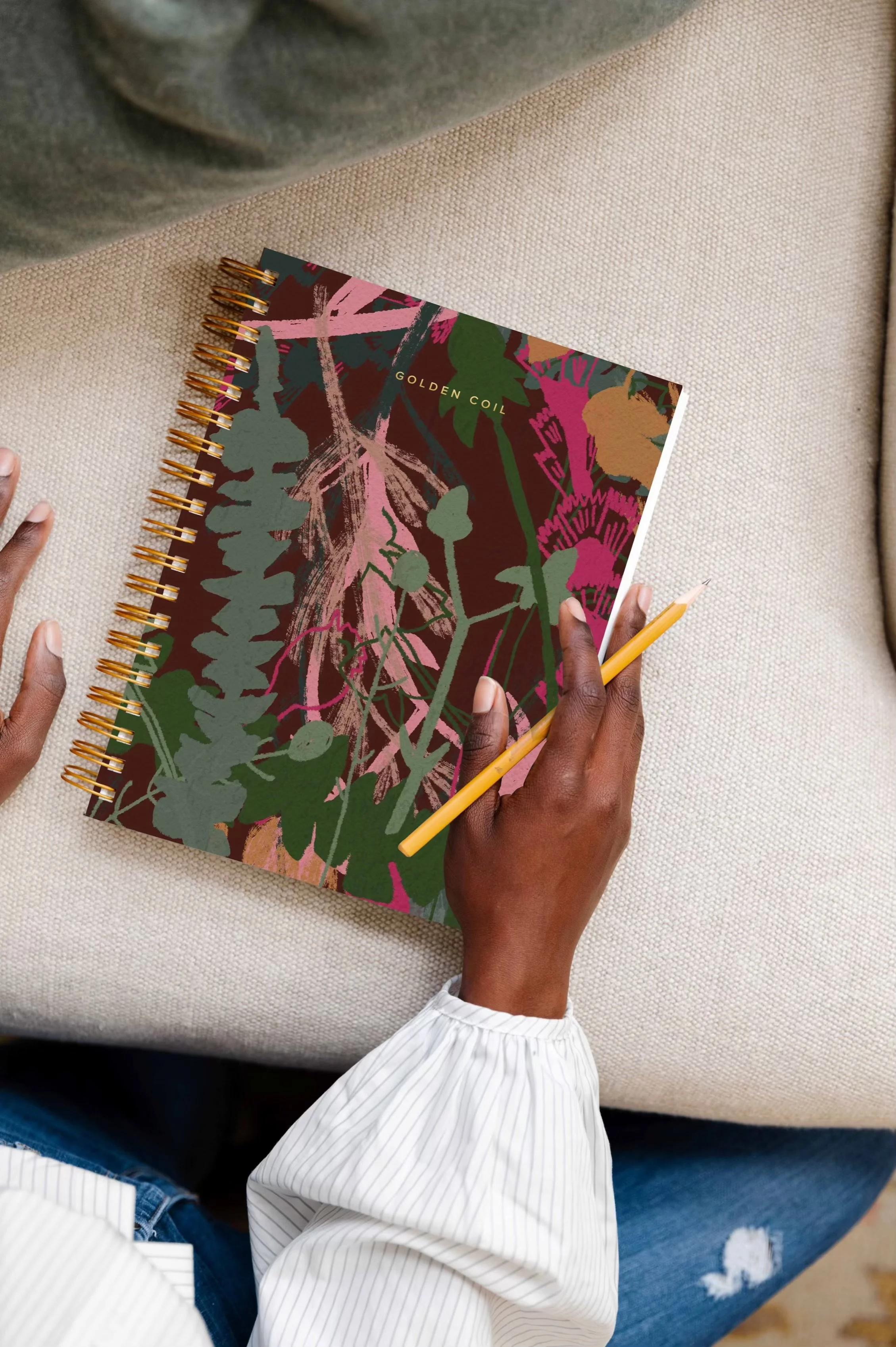 Person holding a yellow pencil above a colorful sketchbook with botanical illustrations and the words "GOLDEN COIL" on the cover, sitting on a beige couch.