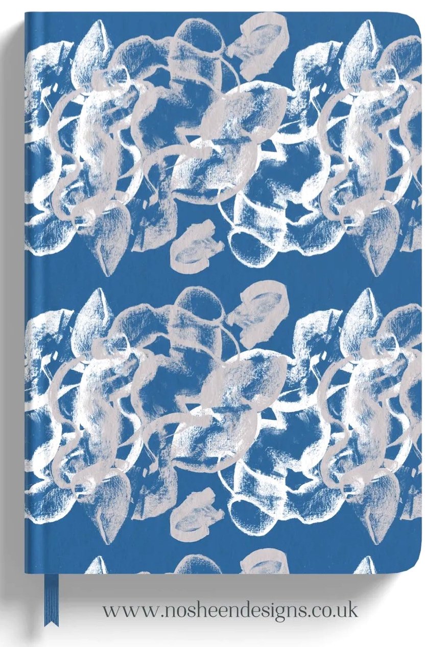 A blue notebook with a white abstract floral pattern on the cover and a ribbon bookmark at the bottom. The website www.nosheendesigns.co.uk is printed at the bottom.