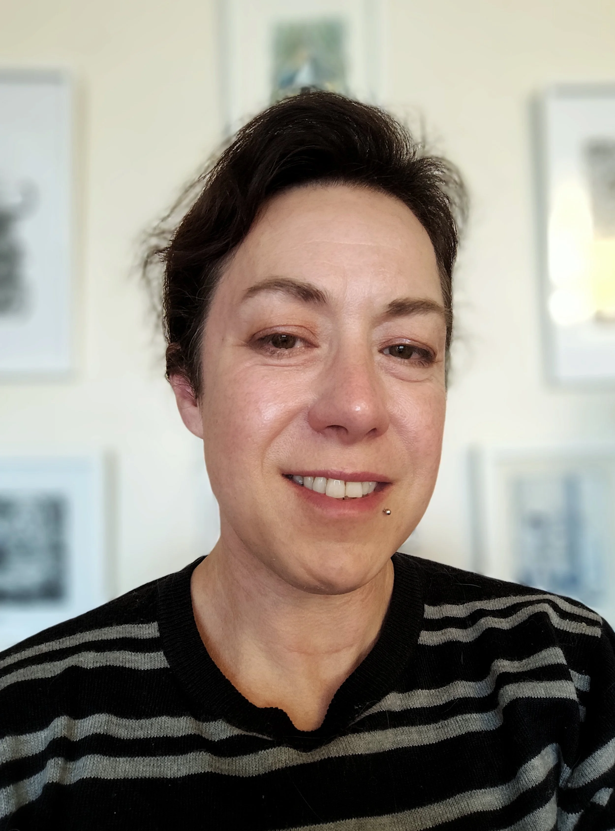 Headshot of author CL Hellisen, smiling at the camera. They have short dark hair, brown eyes, and wear a black jumper with grey stripes.