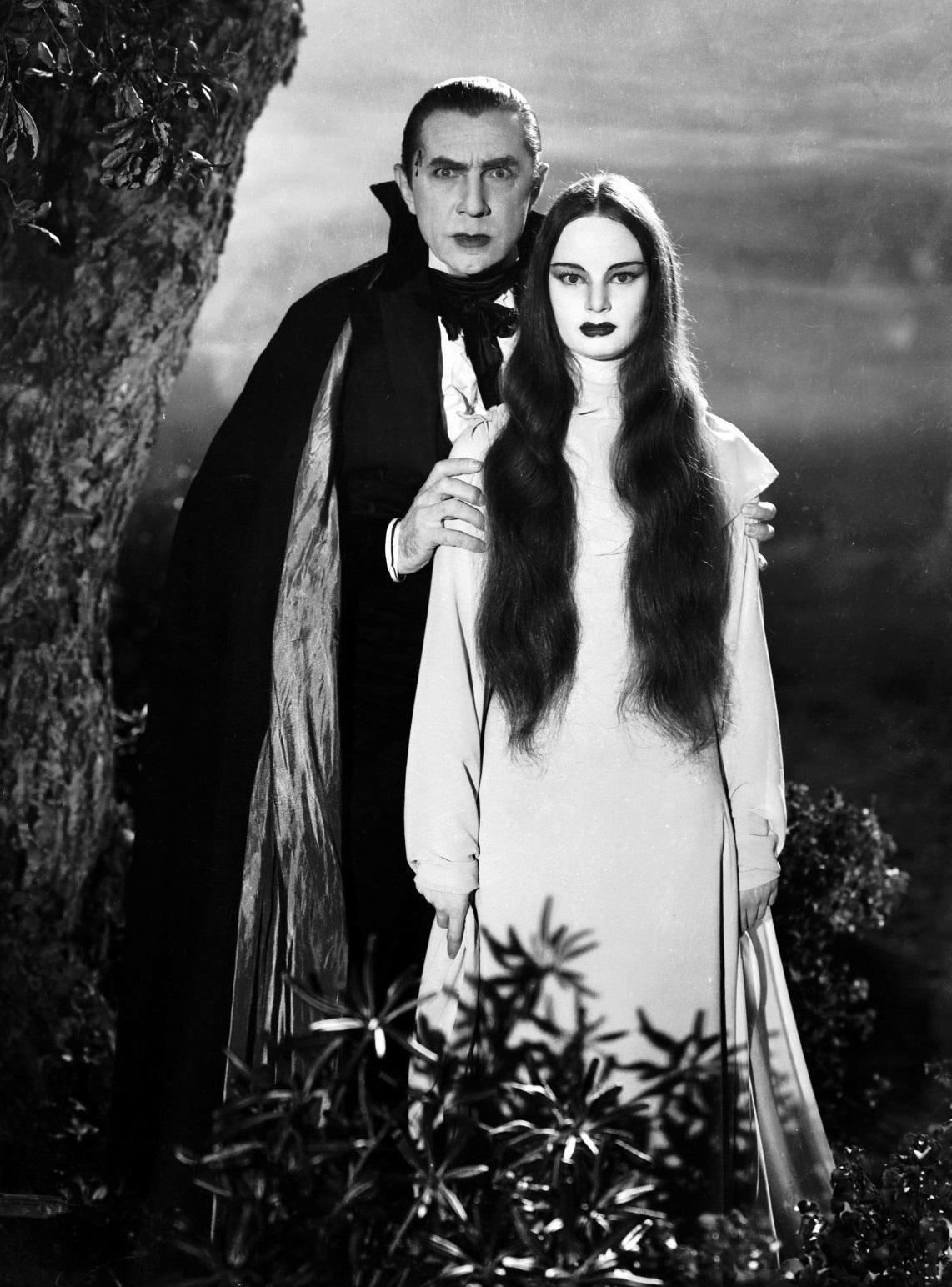 Bela Lugosi as Count Mora stands beside a tree, his hands clasped on the shoulders of Luna (Carol Borland). He is in typical vampire garb, she in a white dress.