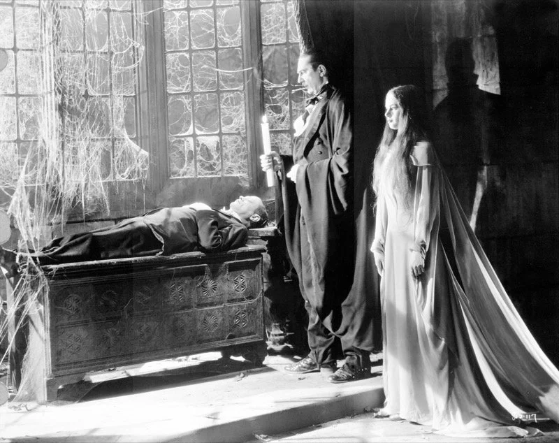 A male vampire stands over a dresser/cabinet with a man stretched out on it, his hands over his chest. Beside the vampire is a female vampire, in a white dress, with long dark hair and white/grey streaks. Image in B&W. Room is old, covered in cobwebs