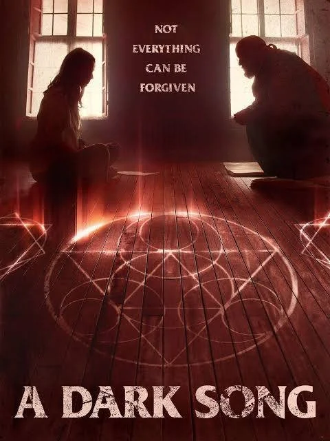 A man and woman sit in the background, near a ritual circle with different symbols. Both are in front of windows. The circle is drawn on a wooden floor, lit up in places.