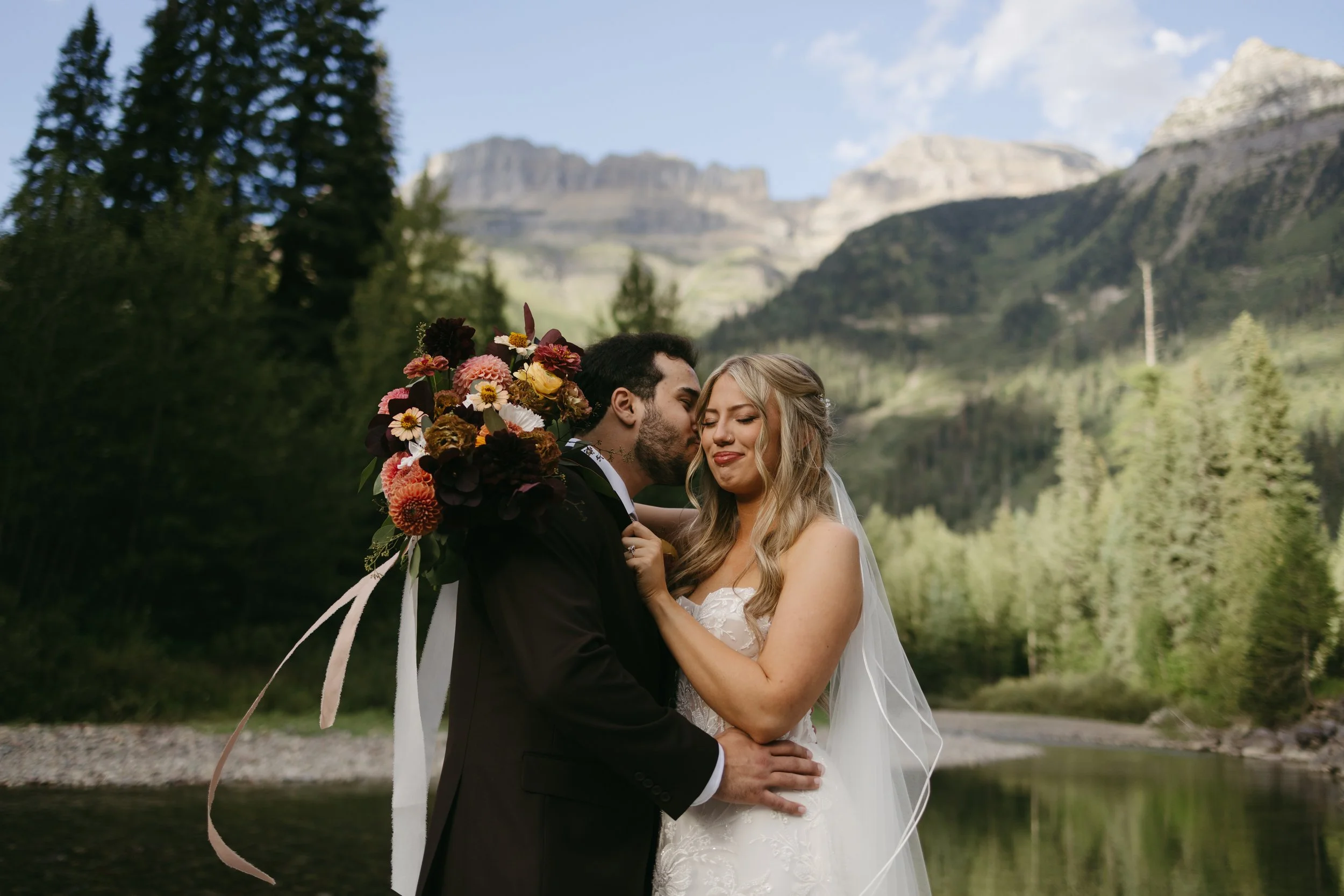 a bride and groom standing in front of the mountains with the groom giving his bride a kiss on the cheek