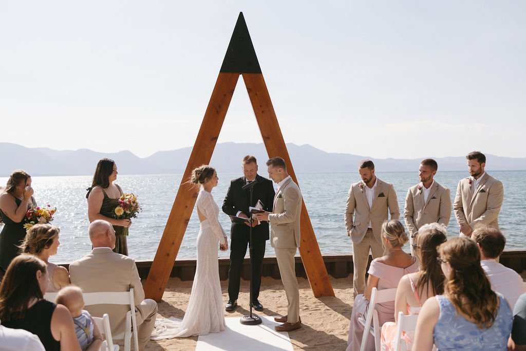 A beach wedding ceremony with a couple exchanging vows under a wooden triangular arch with mountains and water in the background. Guests seated on white chairs observe the ceremony.