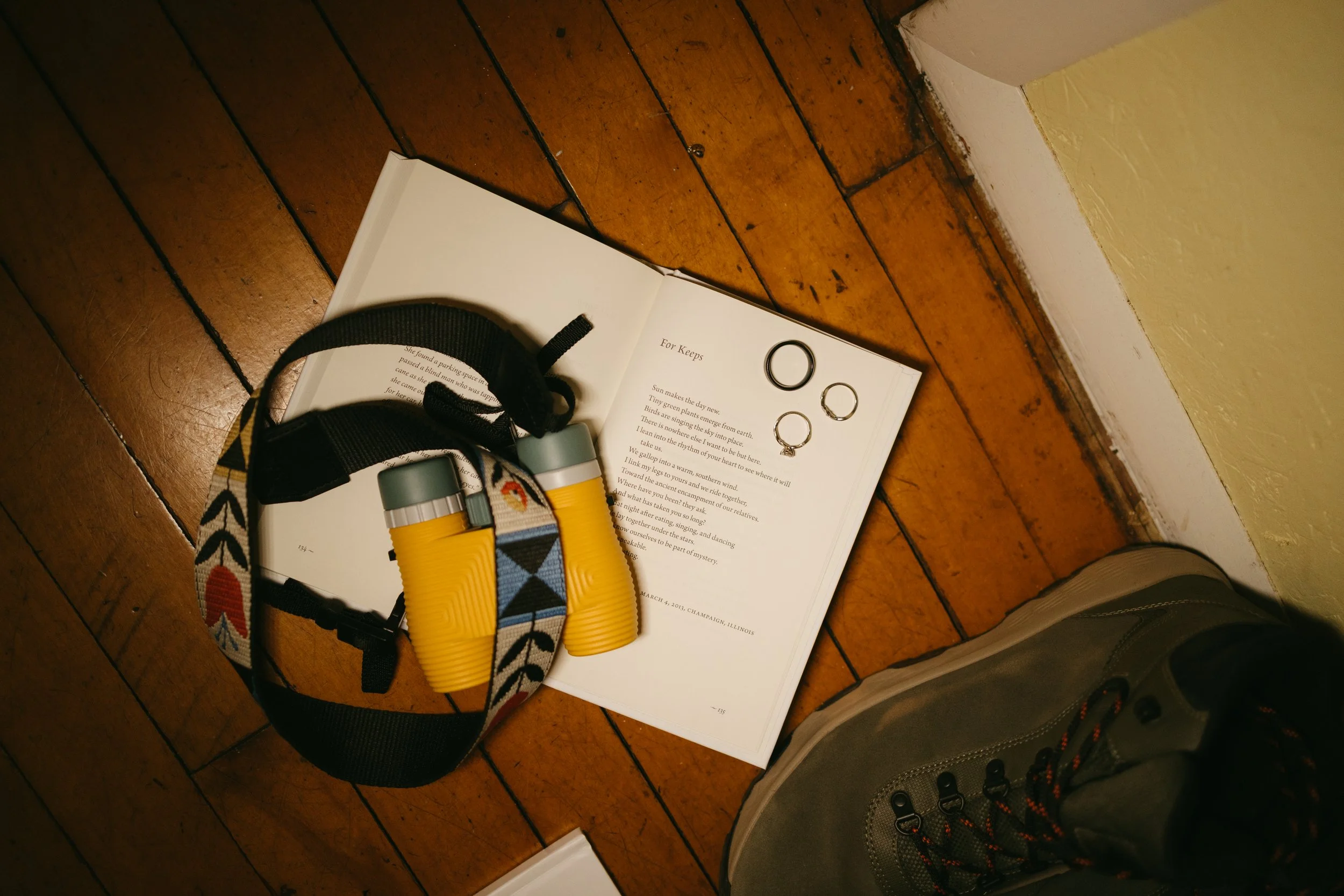 Open book with rings on the page, a pair of yellow binoculars with a strap featuring a colorful chevron pattern, and a person's foot wearing a gray hiking boot on a wooden floor.