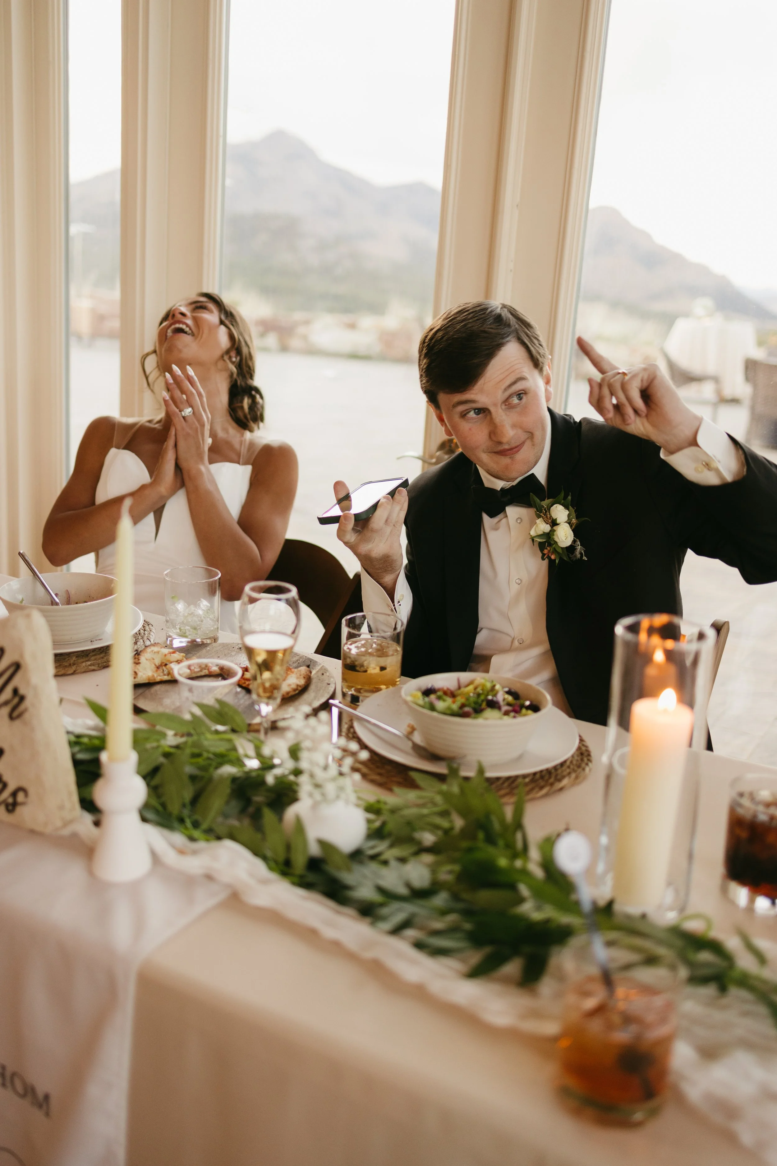 A man and woman in formal attire celebrating at their wedding reception, with mountains visible through large windows in the background, surrounded by food, drinks, and candles.