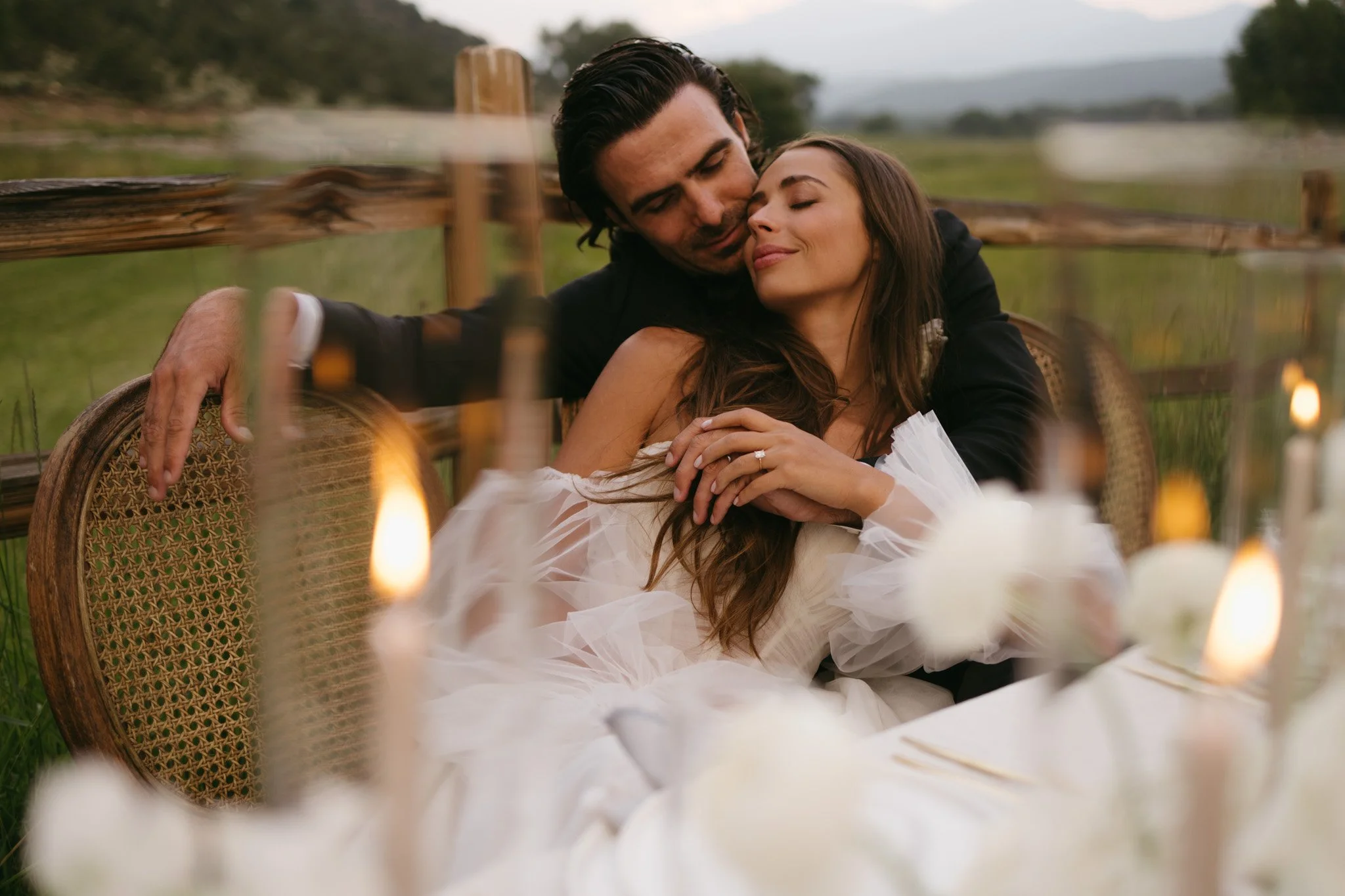 A couple embracing at a rustic outdoor setting with candles and greenery.