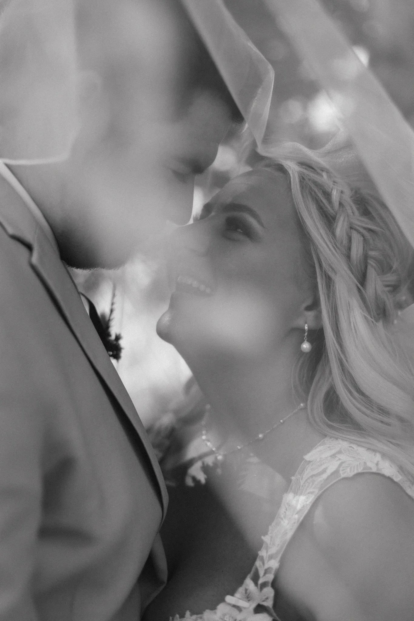 A black-and-white photo of a couple close together, smiling and looking into each other's eyes, under a sheer veil, with the woman wearing earrings and a necklace.