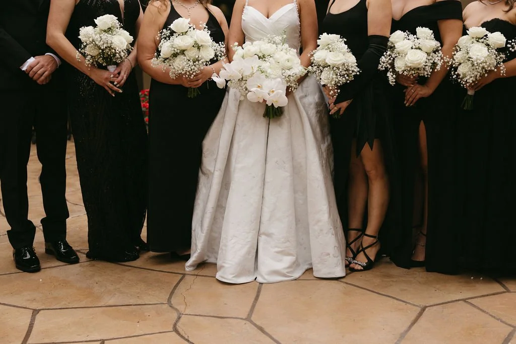 Close-up of a group of women in black dresses holding bouquets of white flowers, centered around a woman in a white wedding gown holding a large bouquet, at a wedding or formal event.