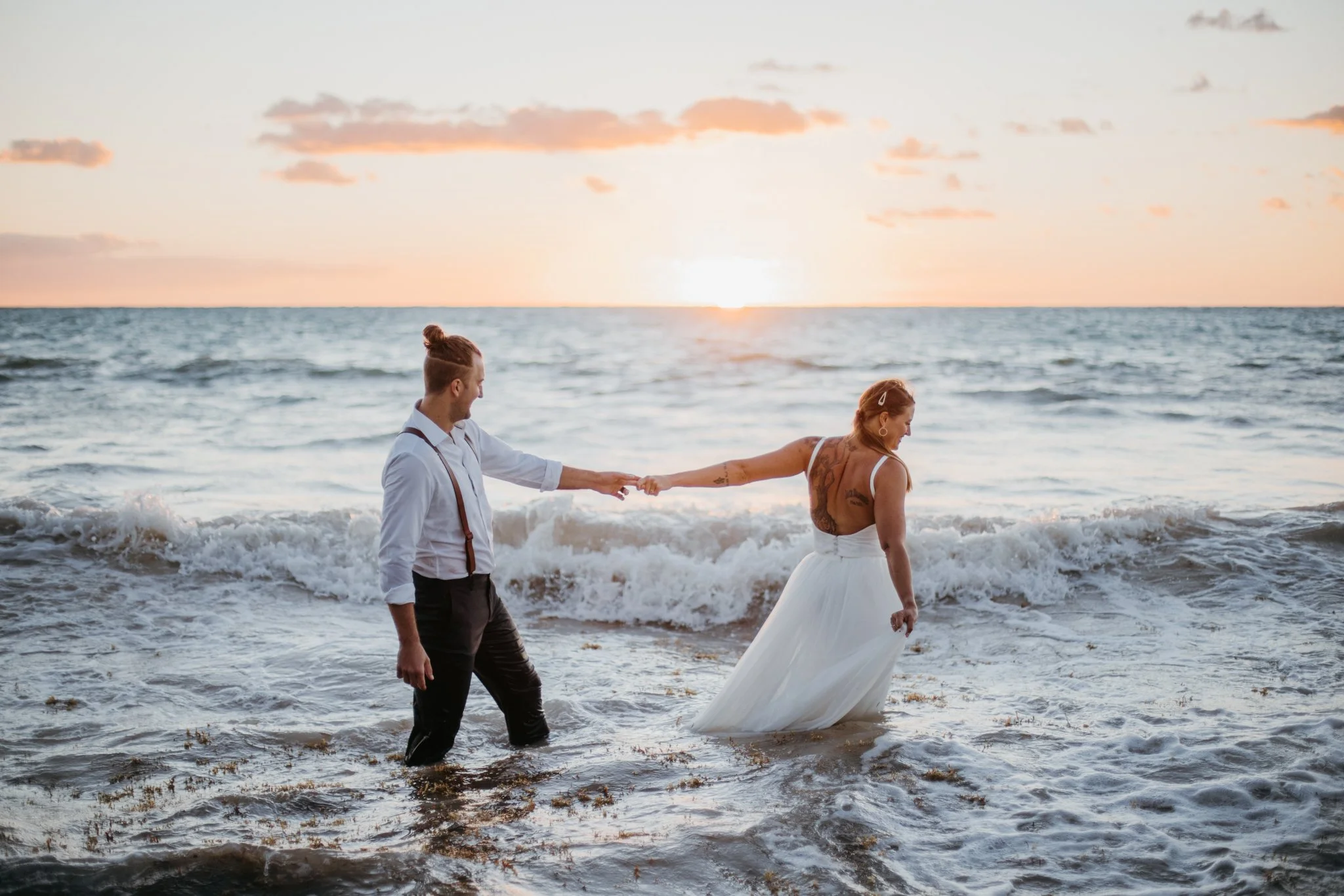 A couple holding hands at the beach during sunset, with the woman in a white dress and the man in a white shirt and suspenders, standing in the water.