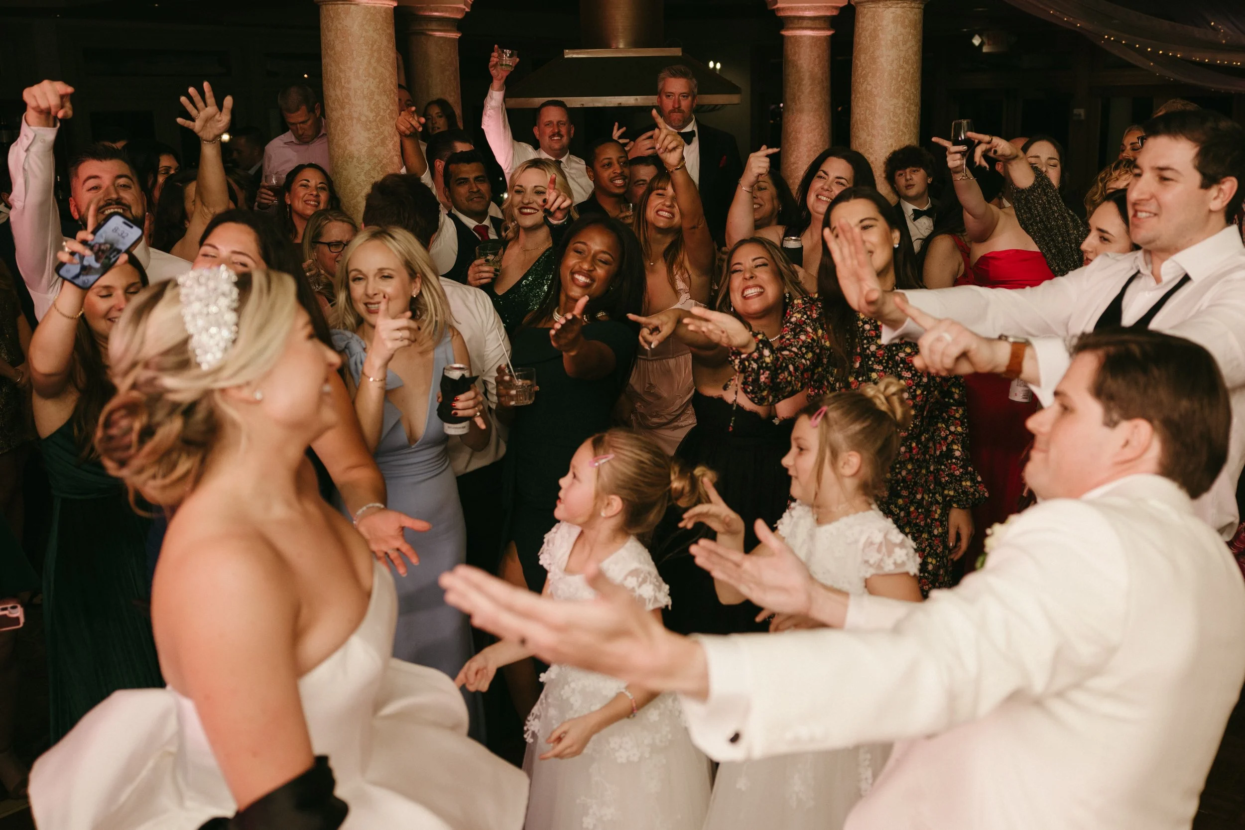 A wedding reception with the bride and groom dancing surrounded by guests cheering and taking photos.