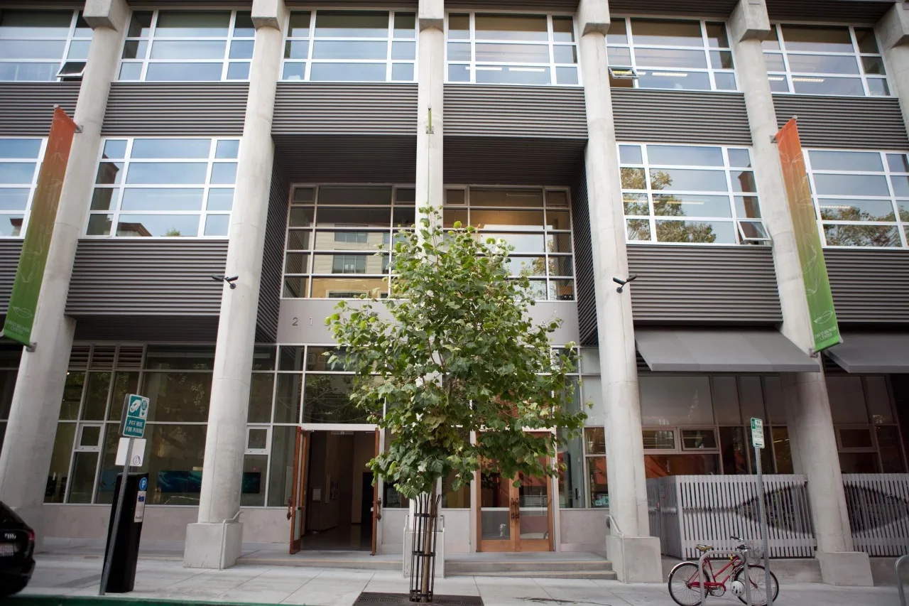 Modern building with large windows, neighborhood street scene with a tree, bike, and parking signs.