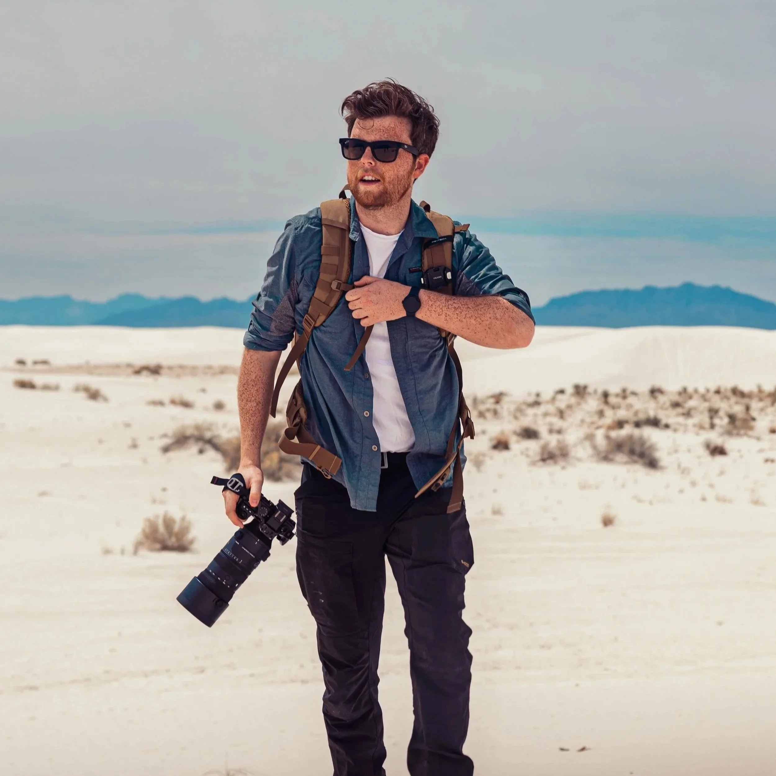 A man with sunglasses and a beard stands outdoors in a snowy area holding a large camera lens, wearing a blue jacket and a backpack.