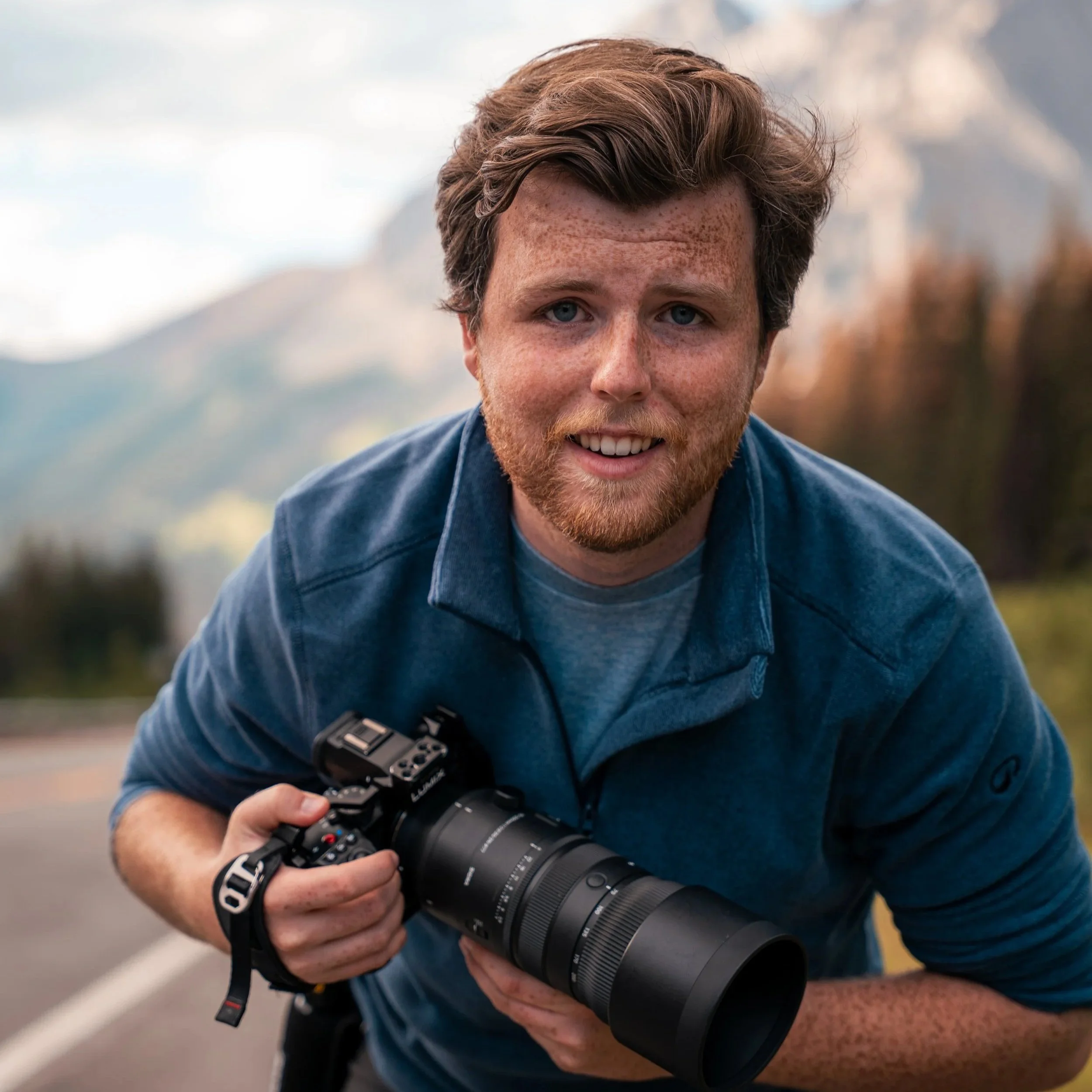 A man with red hair, a beard, and freckles holding a camera with both hands, outdoors in a blurred natural background.