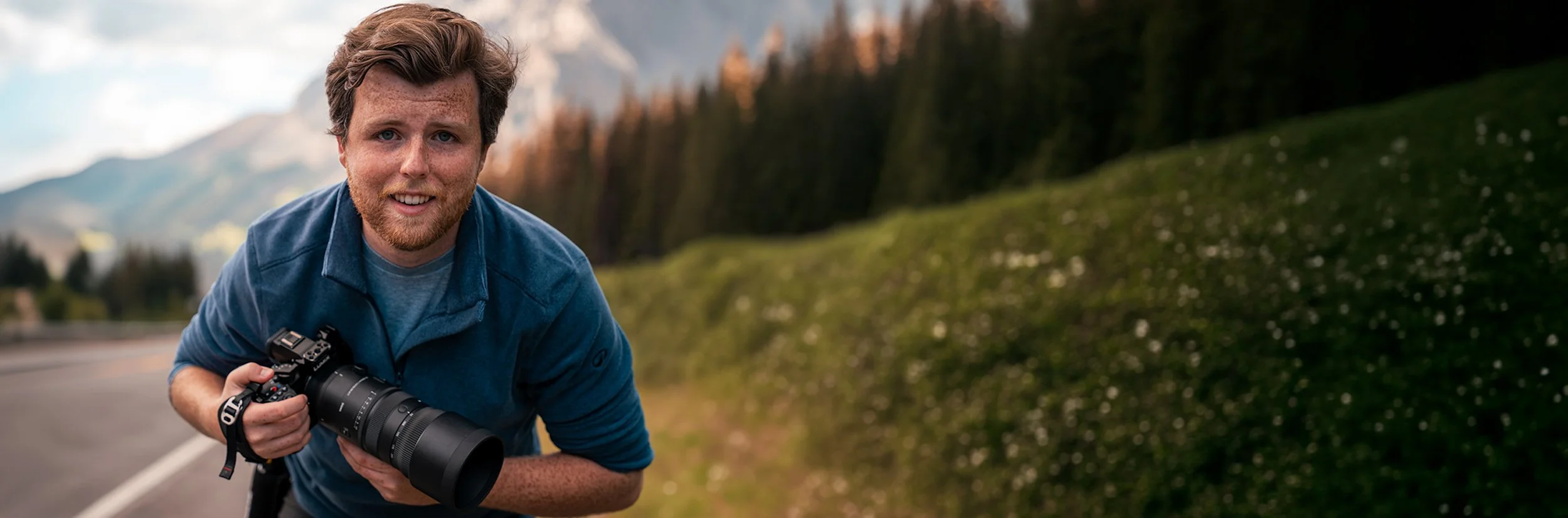 A man with curly brown hair and a beard, wearing a blue jacket and gray shirt, smiling while holding a camera with a large telephoto lens, crouching on the side of a road with mountains and a forested hill in the background.