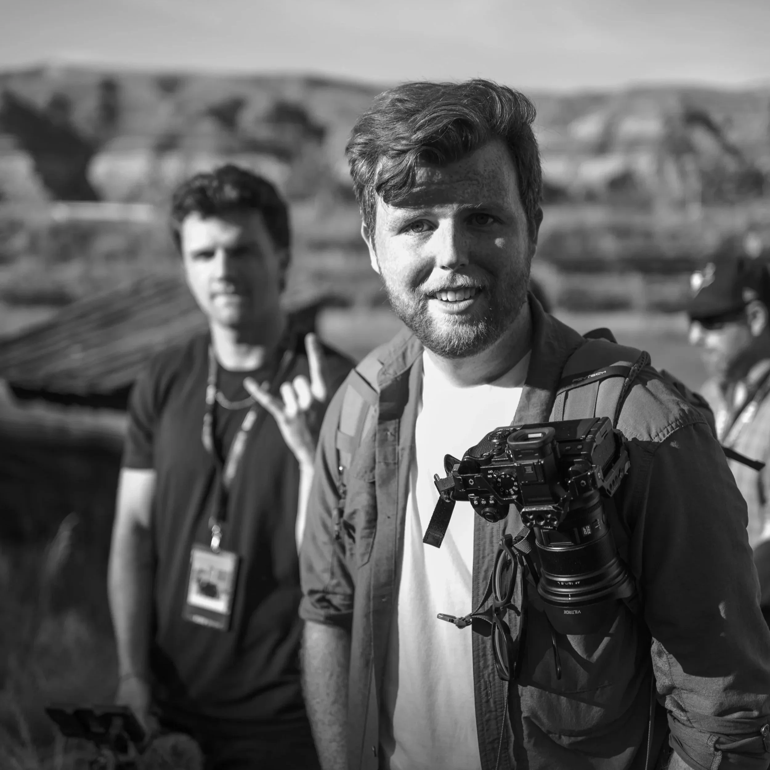 Two young men outdoors in front of a bridge, one in the foreground wearing a jacket and holding a camera, the other blurred in the background. Black and white photograph.