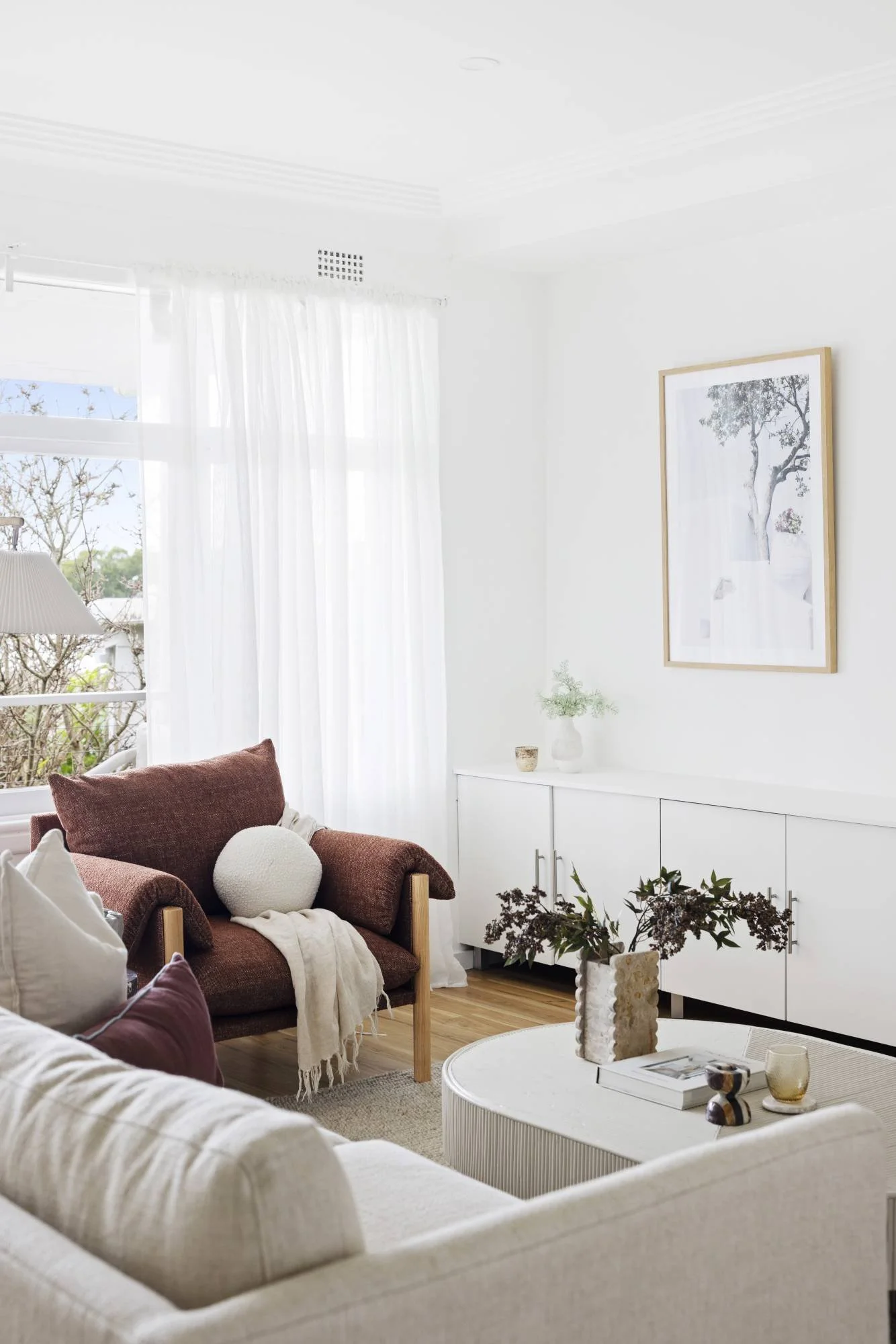 Living room with white walls, large window with sheer curtains, brown armchair with white pillow, white sideboard decorated with plants and framed picture, round coffee table with books and vases, and wooden flooring.