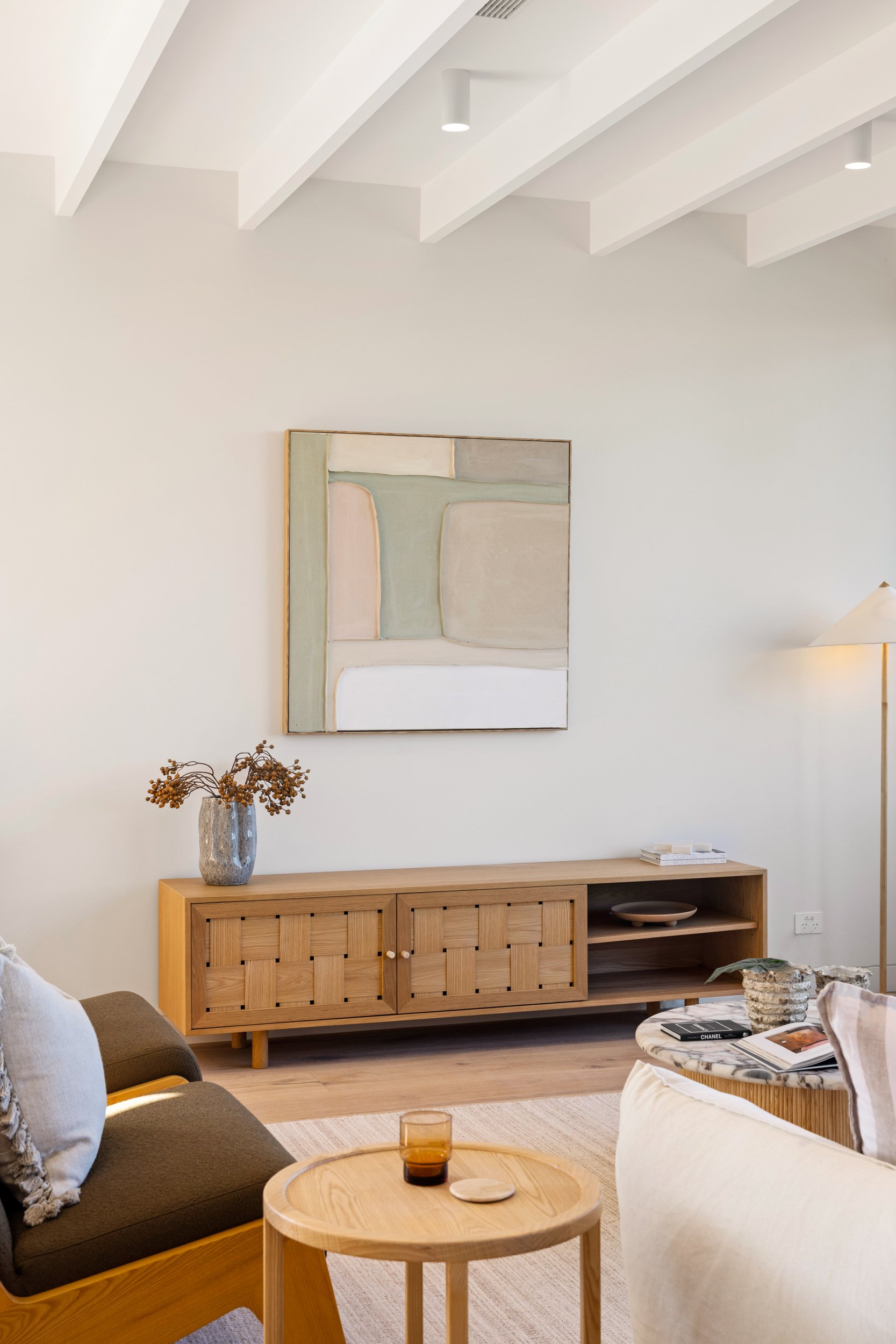 Living room with white walls, modern wood sideboard, abstract wall art, a grey sofa, a round marble coffee table, books, a vase with dried flowers, and decorative items.