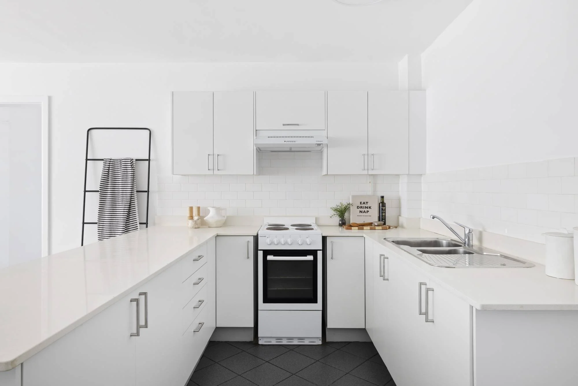 A minimalistic white kitchen with white cabinets, a white stove, and a white countertop. There is a black towel rack with a striped towel, and a sign that says 'Eat Drink Nap' on the counter. The kitchen has a sink with a faucet and some small decora