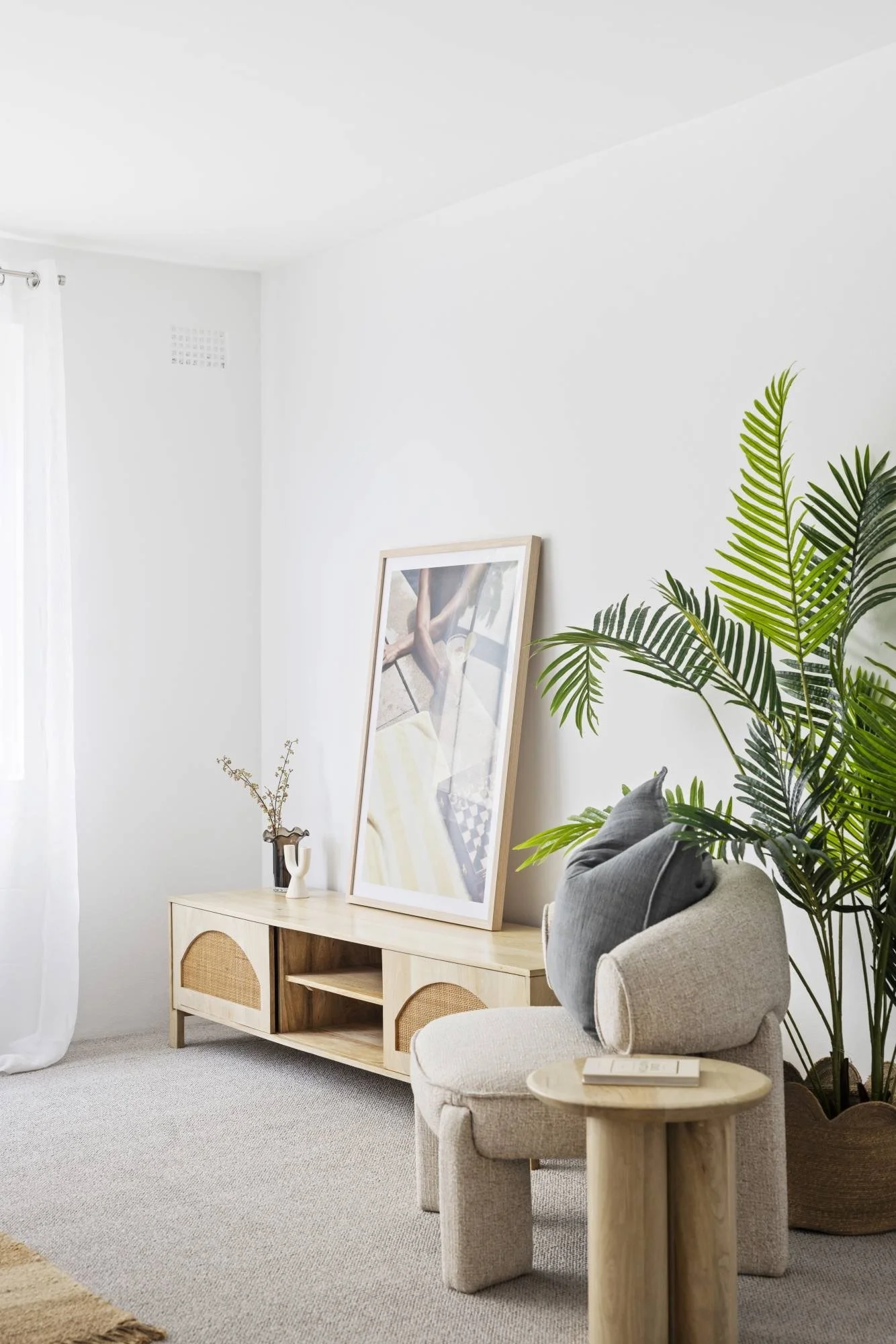 A cozy living room corner with a light beige armchair with a dark gray pillow, next to a small round wooden side table with a book. A large green plant in a woven basket is beside the chair. A wooden console table against the white wall holds a frame