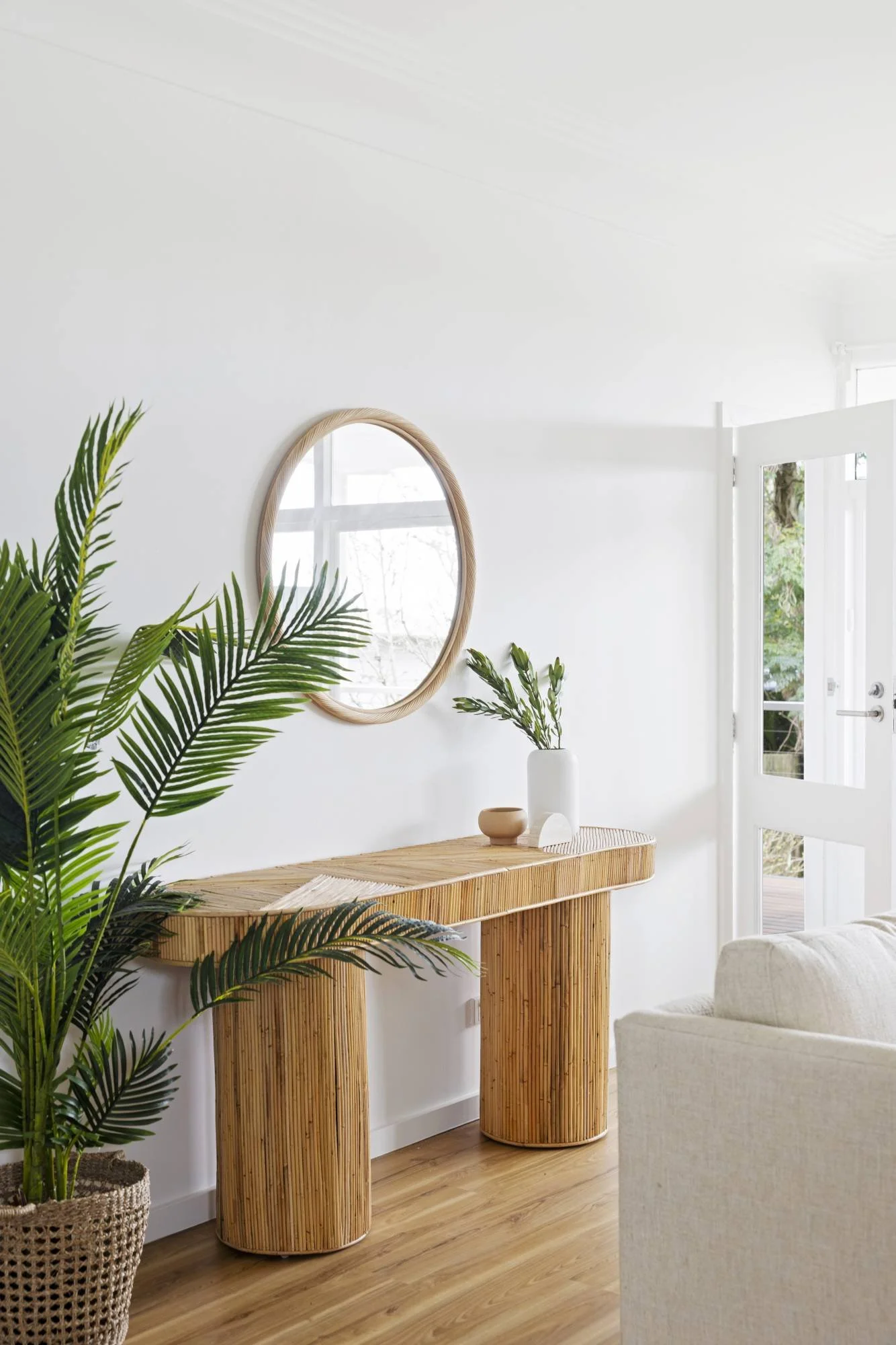A bright, minimalist living space with vertical wooden furniture, green potted plants, a round mirror, and an open door with a view of trees outside.
