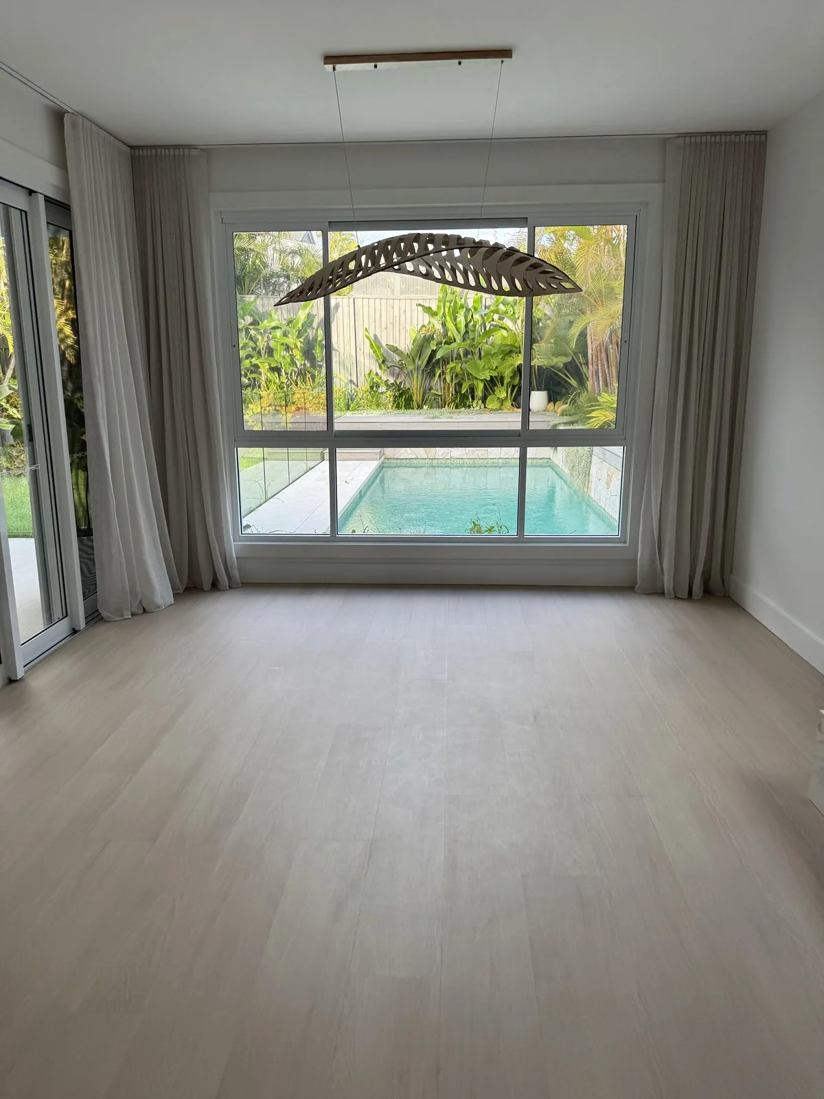 Empty room with large window overlooking a backyard pool and lush green plants.