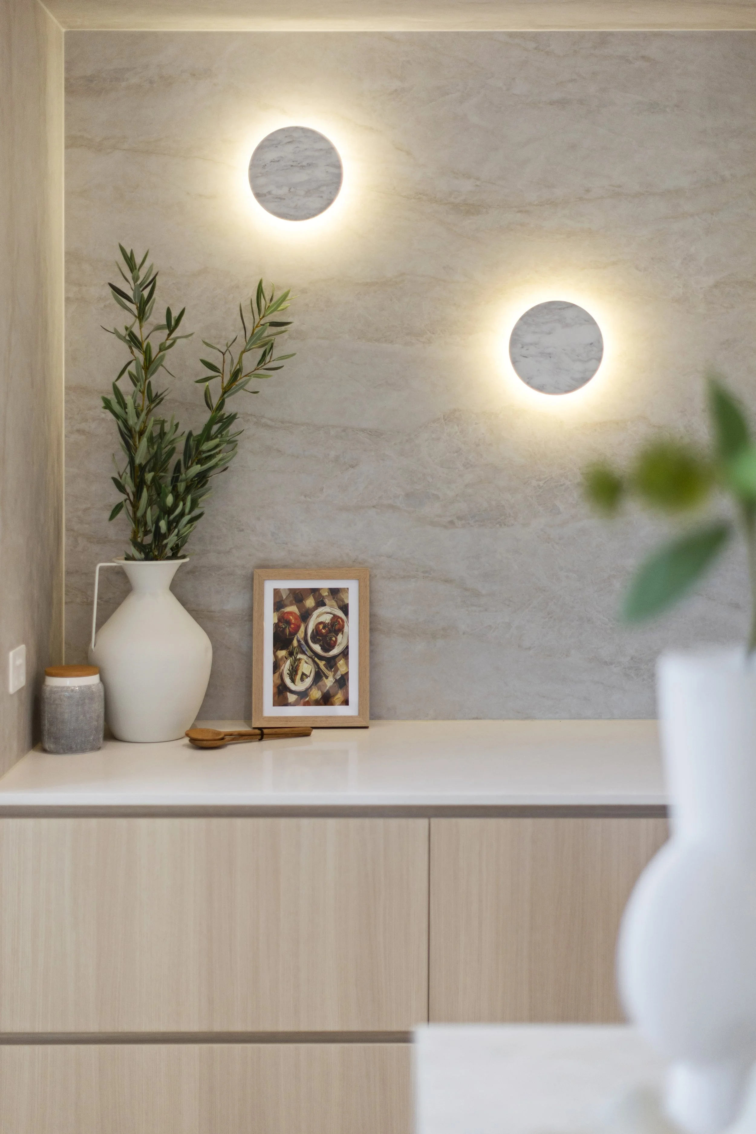 Decorative wall with two round, backlit marble-textured wall sconces, a potted olive branch in a white vase, a framed picture of apples, and a small container on a white countertop.