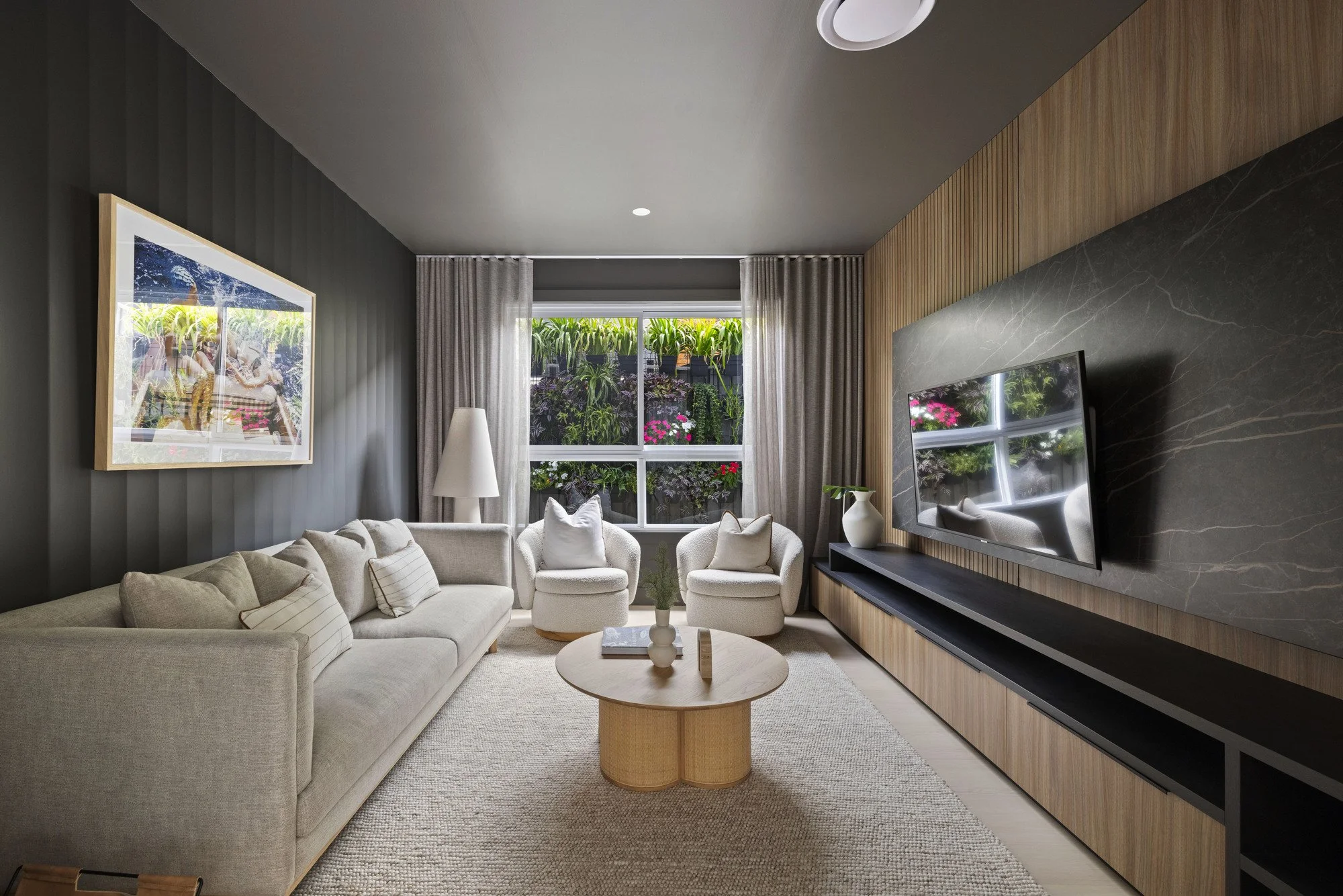 Living room with beige sofa and two armchairs, a round coffee table, a framed picture on the gray wall, a TV on a wood panel wall, and large windows with curtains showing greenery outside.