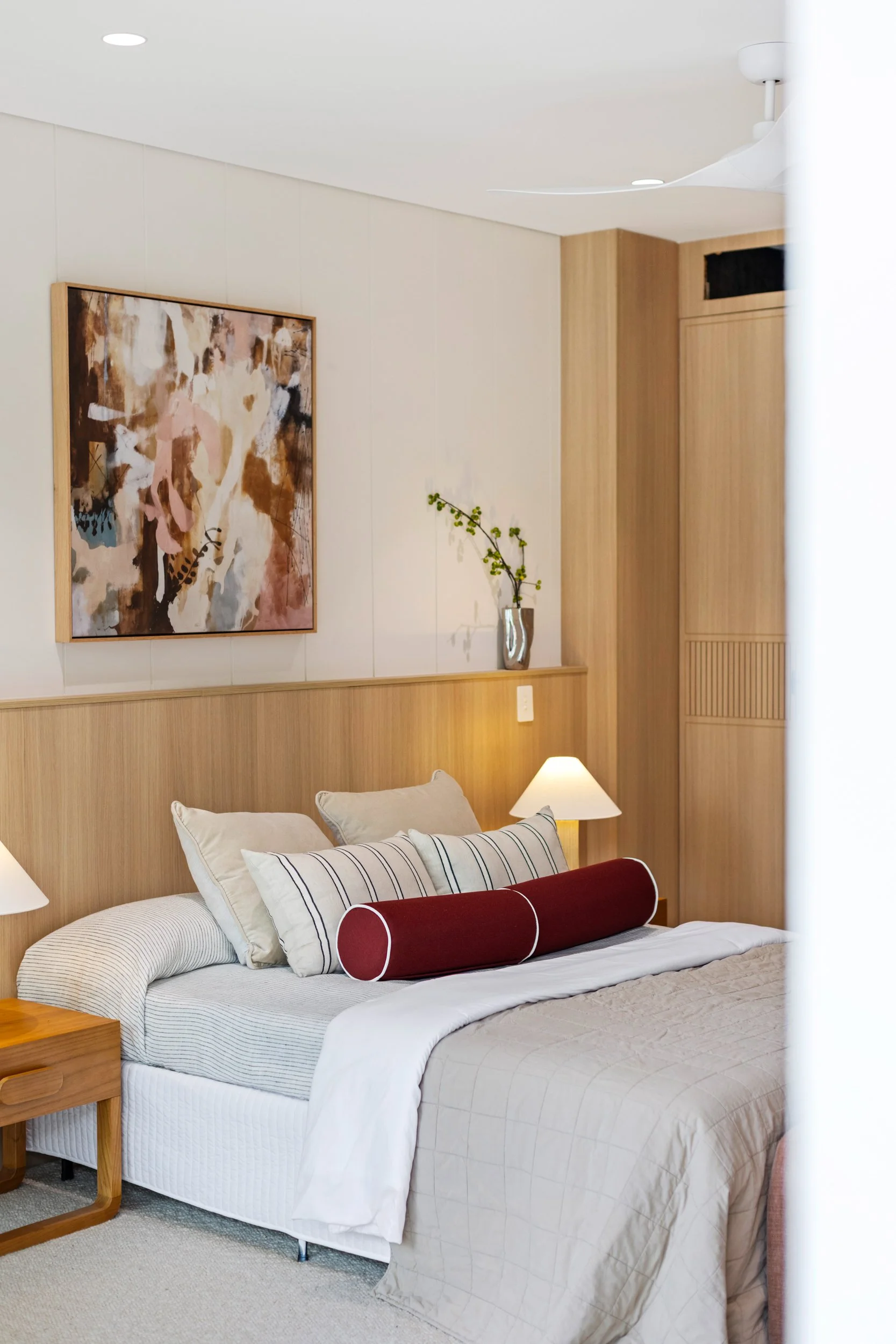 A neatly made bed with white and gray bedding, multiple pillows with striped and plain pillowcases, a red cylindrical bolster, a small wooden bedside table, a white bedside lamp, wall art with an abstract painting, a tall vase with a single branch of green berries, a wooden headboard with paneling, and a ceiling fan in a modern bedroom with beige carpet and wooden panel accents.
