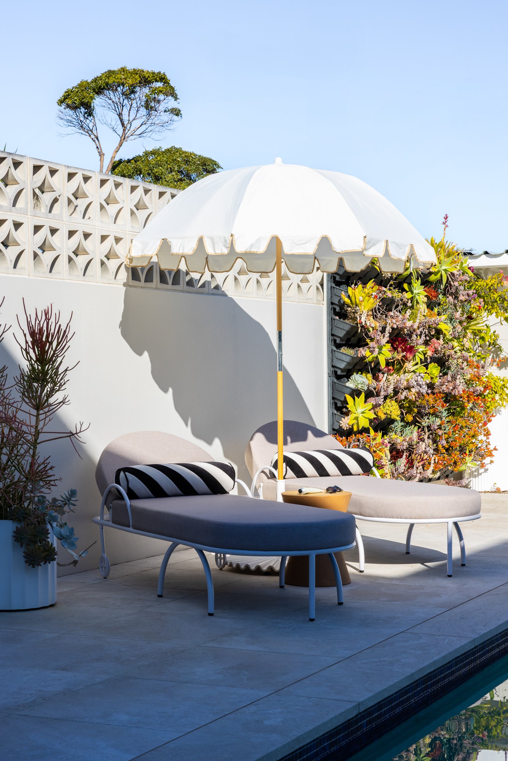 Sunlit outdoor patio with modern lounge chairs, a large white umbrella, and a vibrant vertical garden of flowers against a white wall, overlooking a pool.