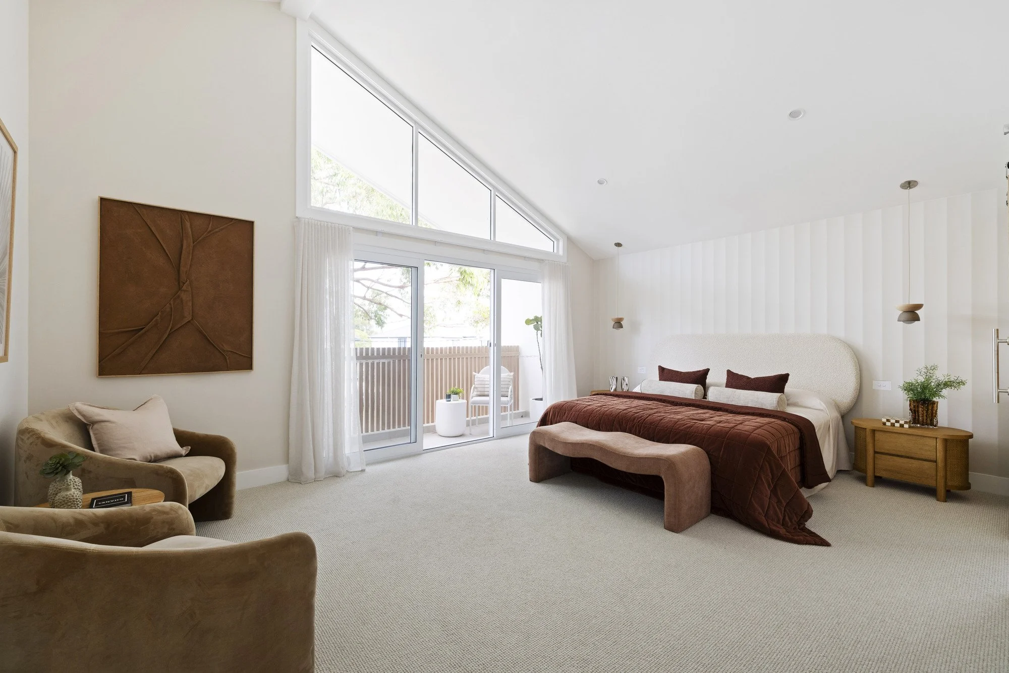 A spacious bedroom with a high sloped ceiling, large windows, and a balcony door letting in natural light. The room features a bed with a brown quilt, matching pillows, two pendant lights, a small wooden nightstand with a plant, and a cozy beige chair with a pillow. There is artistic wall decor and white curtains framing the windows.