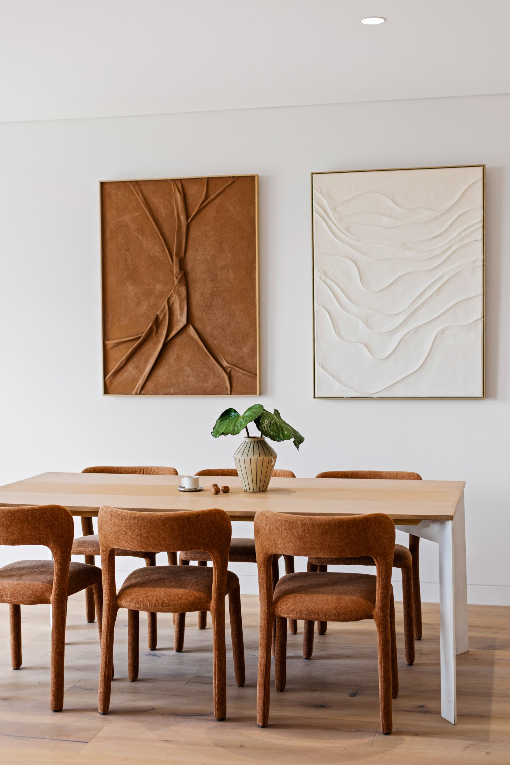 A modern dining room featuring a long wooden table surrounded by multiple orange upholstered chairs. On the table, there is a decorative vase with green leaves, a cup and saucer, and some small objects. The wall behind has two large abstract art piec